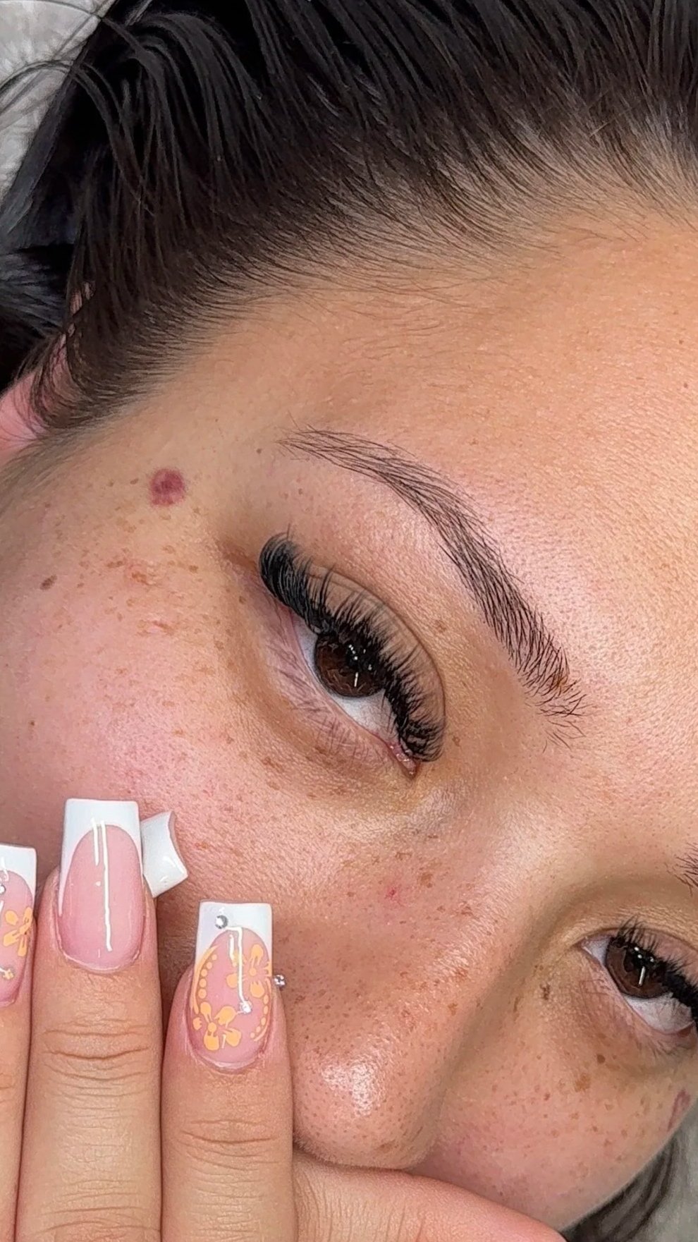 Close-up of a woman's face showing freckles, with her hand near her cheek, having floral nail art and long eyelashes.