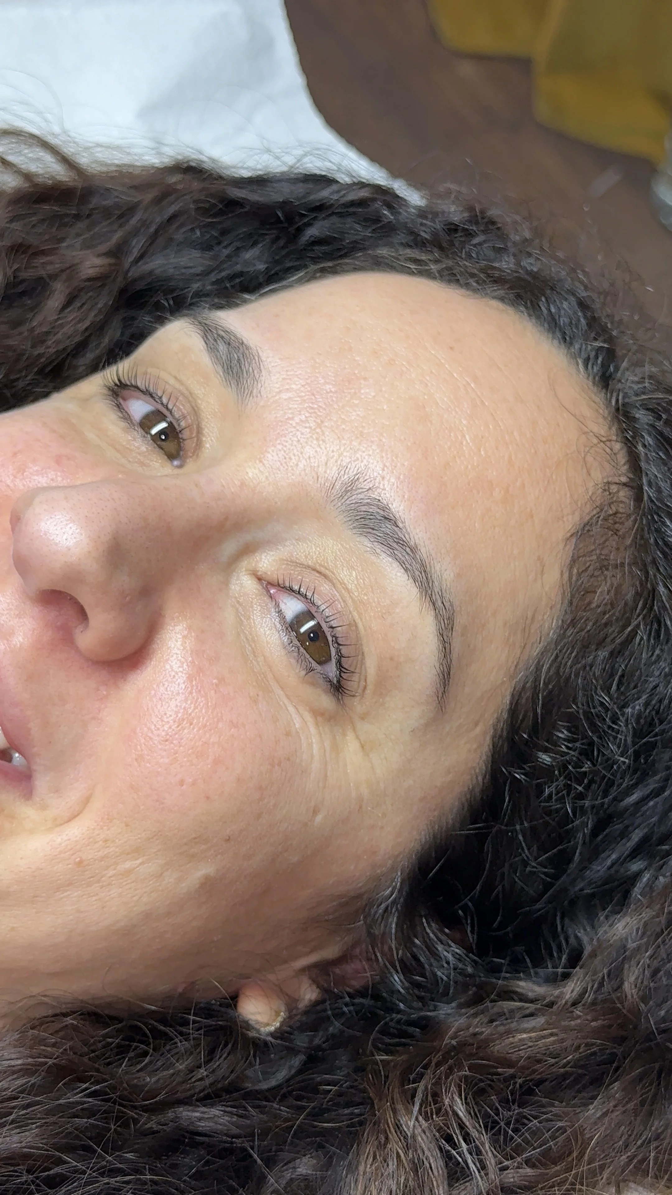 Close-up of a woman's face with curly dark hair, brown eyes, and natural makeup, lying down.