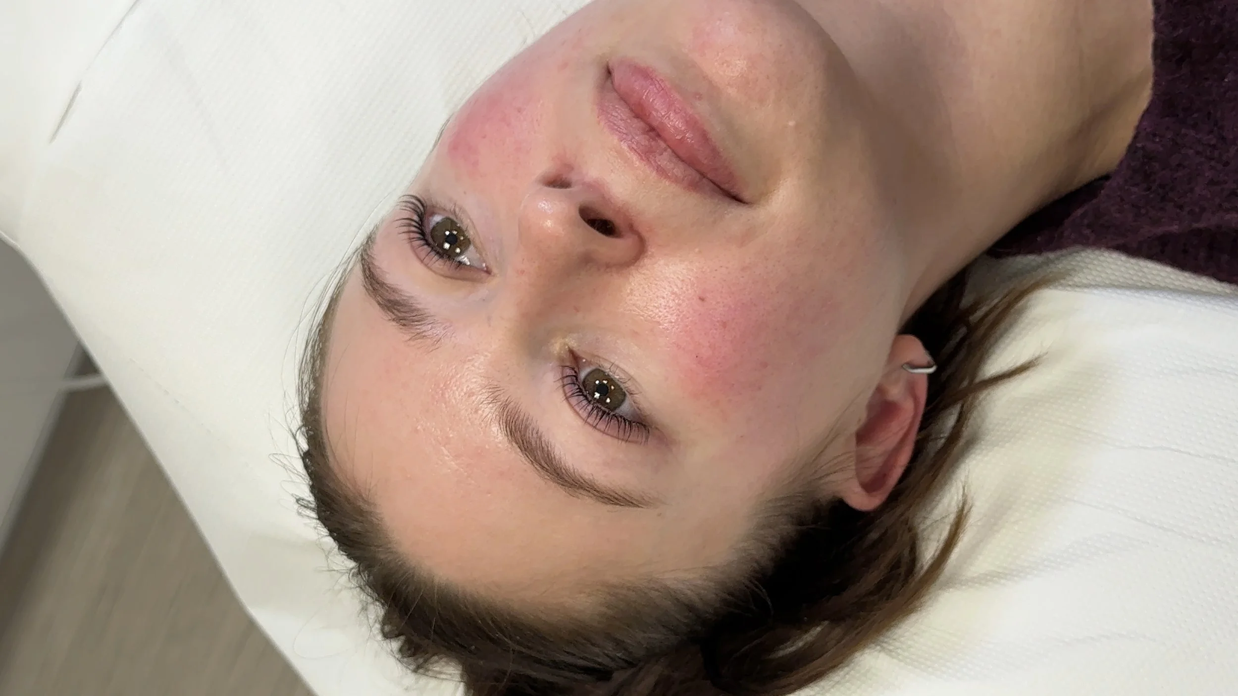 A woman with light skin and brown hair lies on a white surface, gazing up with a relaxed expression.