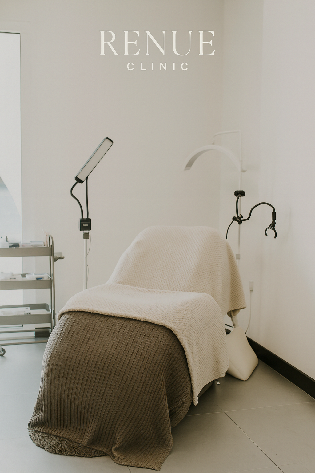 Treatment bed at RENEW Clinic with beige and brown blankets, medical equipment and a sconce light on the wall in a clean, minimalist room.