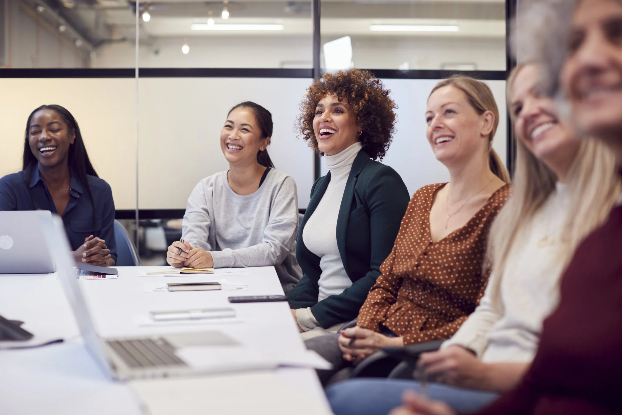 A diverse group of women sitting at a conference table, smiling and laughing during a meeting, with laptops and notepads in front of them.