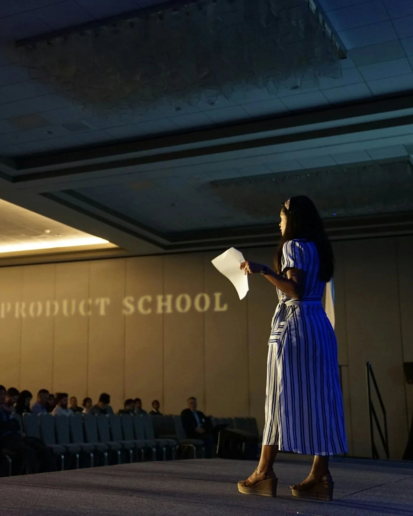 A woman standing on a stage holding a paper, giving a presentation at a conference or school event. The background shows the words "PRODUCT SCHOOL" projected on the wall, with an audience seated and watching her.