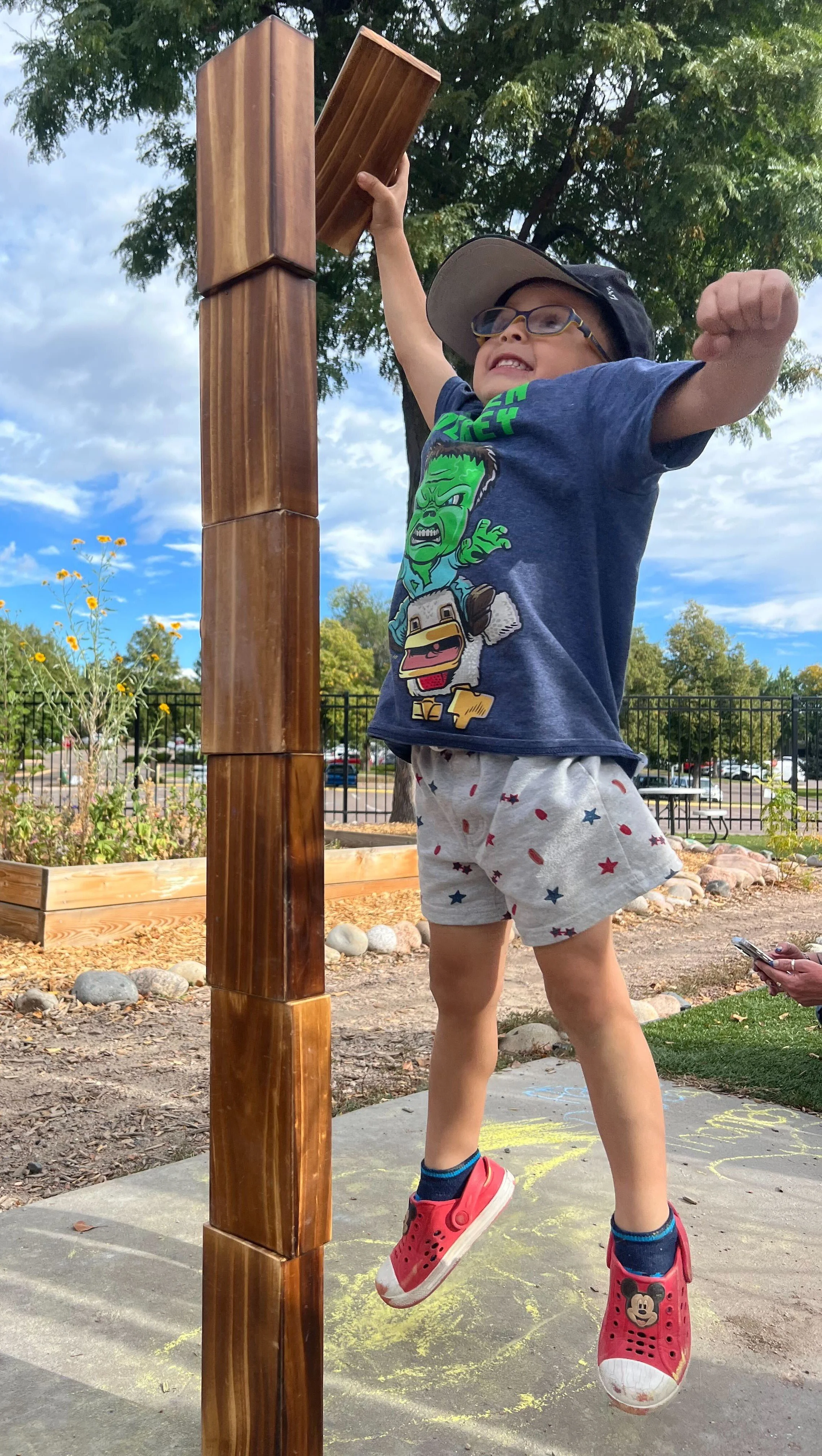 A child jumps to try to stack a block onto a tall block tower outside.
