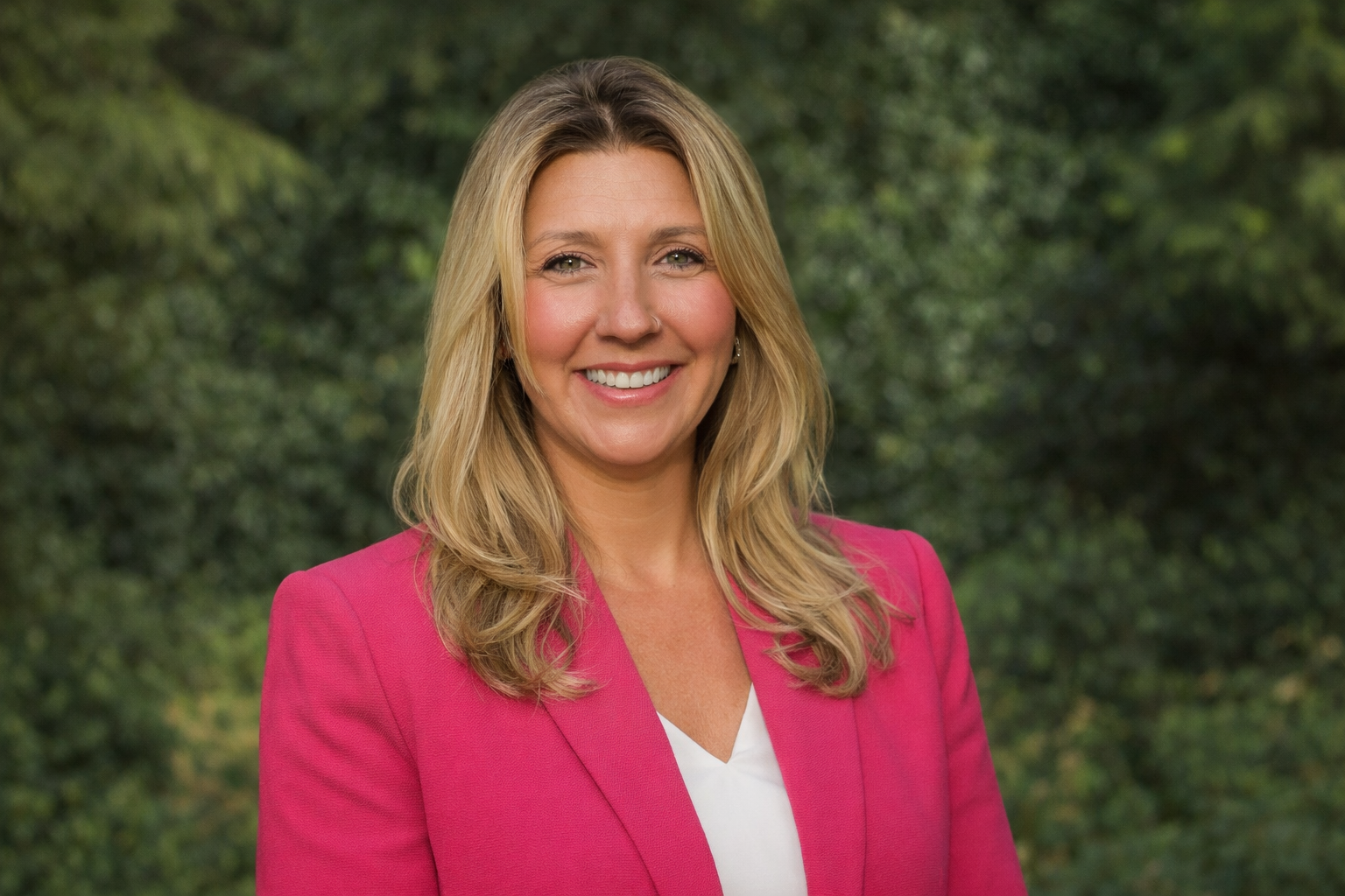 Professional woman with blonde hair smiling outdoors, wearing a pink blazer and white top.