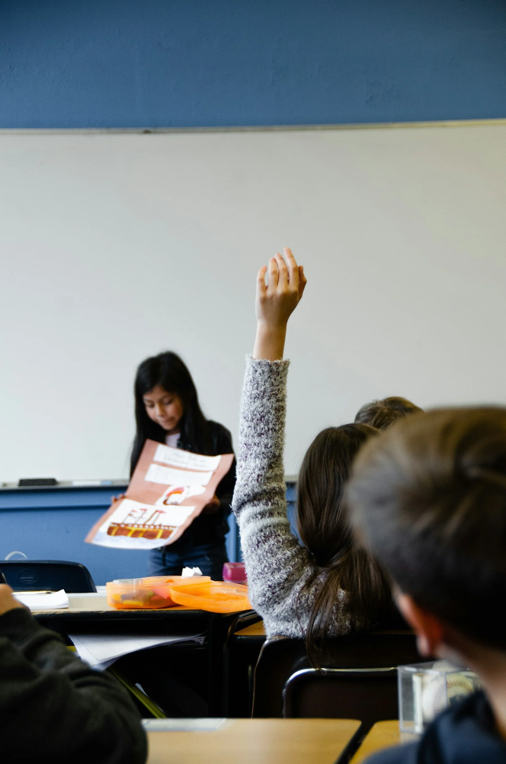 Student presenting in front of class while classmates raise hands for questions.