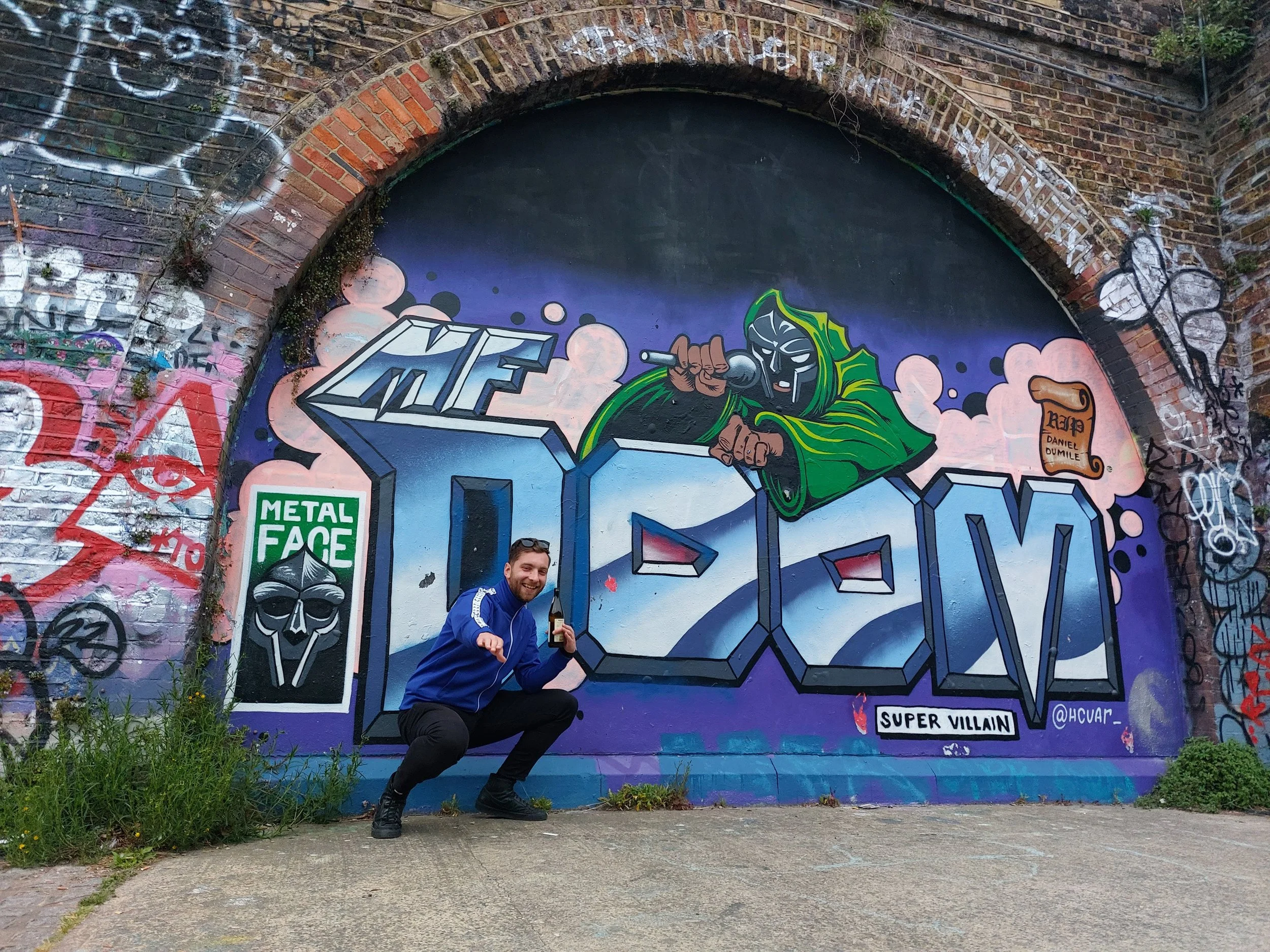 A man crouching and smiling in front of graffiti art that spells "DMV" with a masked figure holding a microphone and a green hooded figure, on a brick wall archway with various tags and artwork.