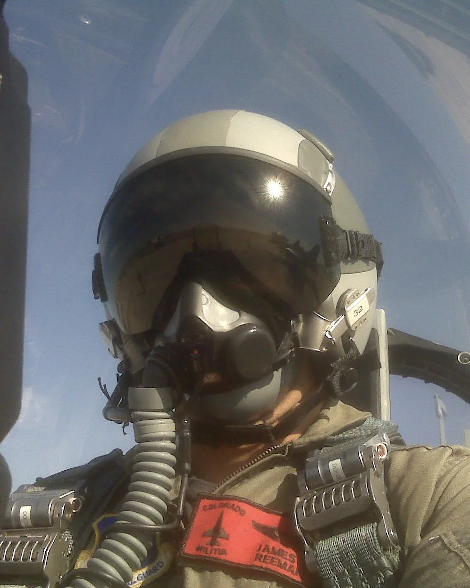 Photo of James Reeman wearing a flightsuit and flight helmet + visor and mask inside of an F-16 Fighter Jet