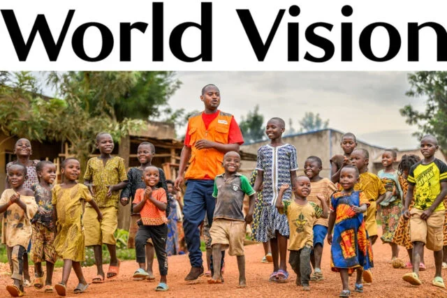 A group of smiling children and an adult man outdoors in a rural setting. The children are wearing colorful clothing, and the man is dressed in an orange vest and dark pants, standing with them.