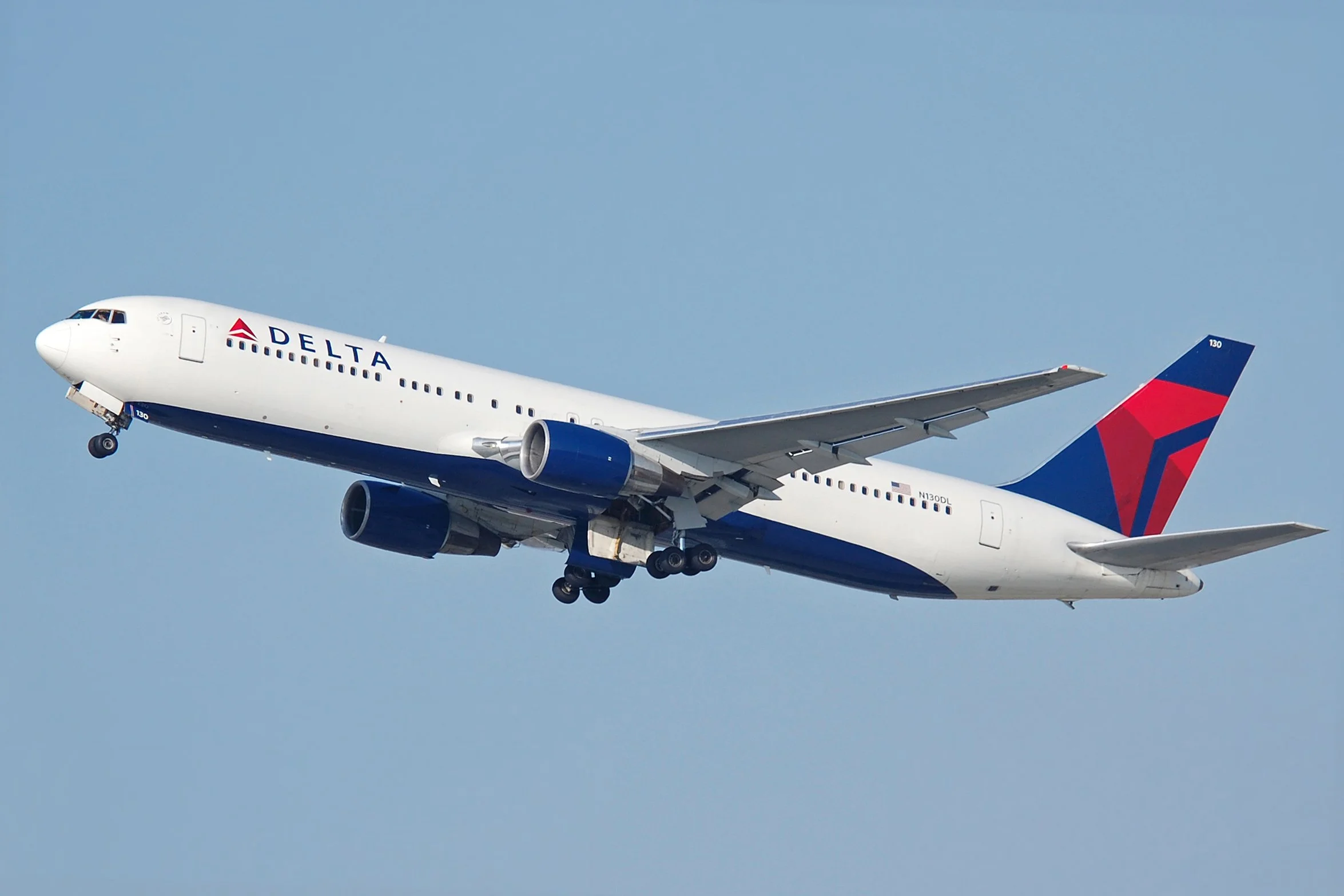 A Delta Airlines commercial airplane flying in the sky during daylight. The airplane is white with blue engines, and has the Delta logo on the tail and the side of the fuselage. The sky is clear with no clouds.