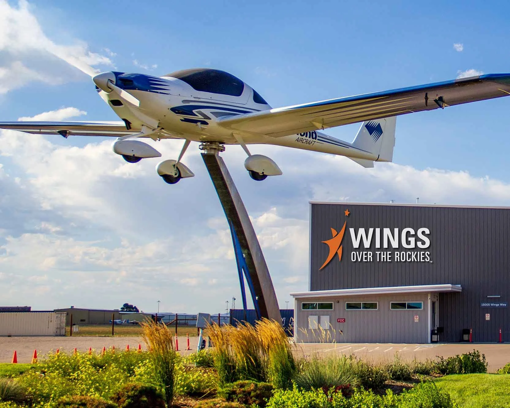 A small white aircraft mounted on a pole outdoors, with a building that has a sign reading 'Wings Over the Rockies' and a partly cloudy sky in the background.