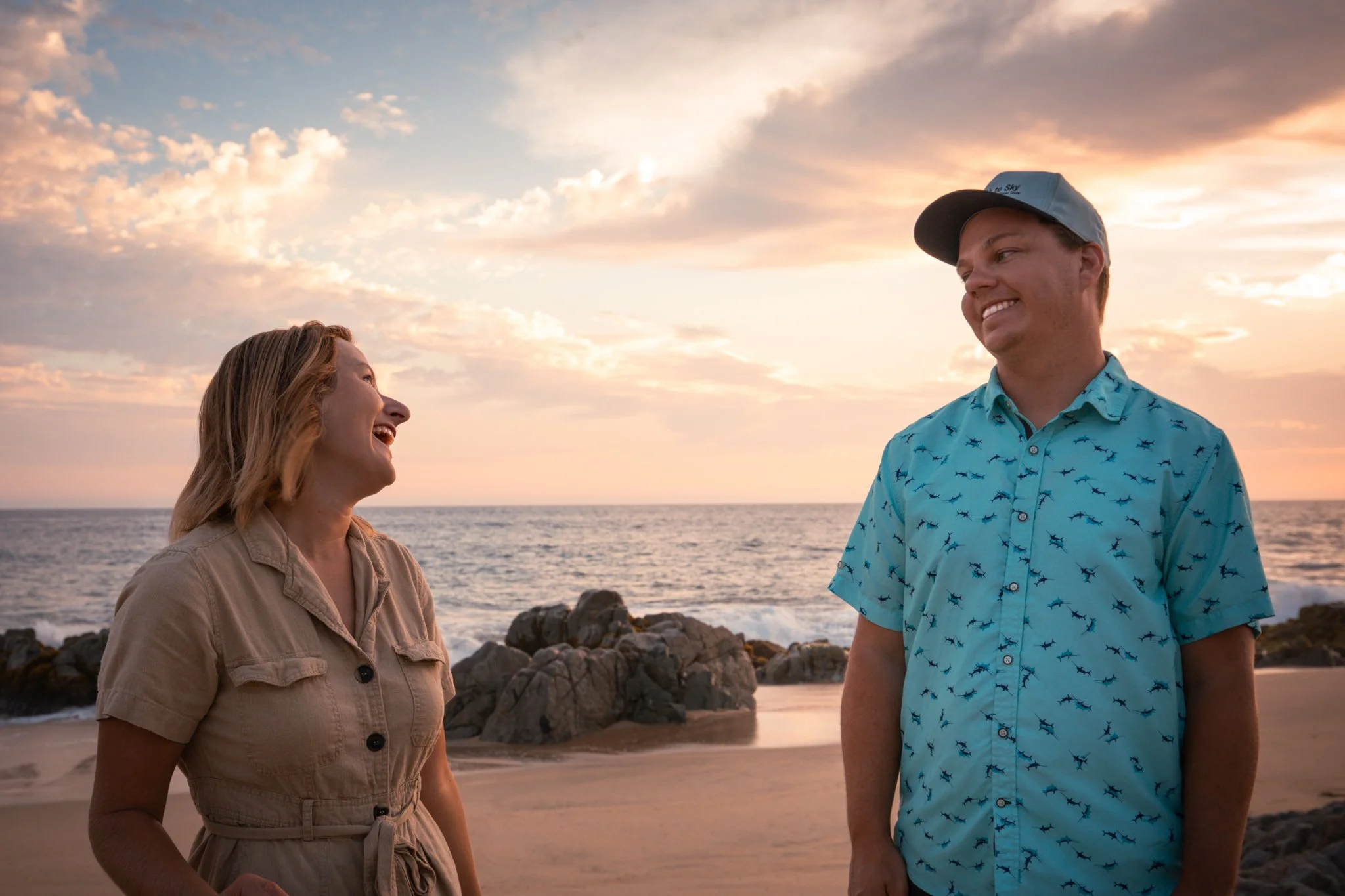 A woman with long blonde hair and a man in a blue shirt with a shark pattern standing on the beach at sunset, smiling and looking at each other.