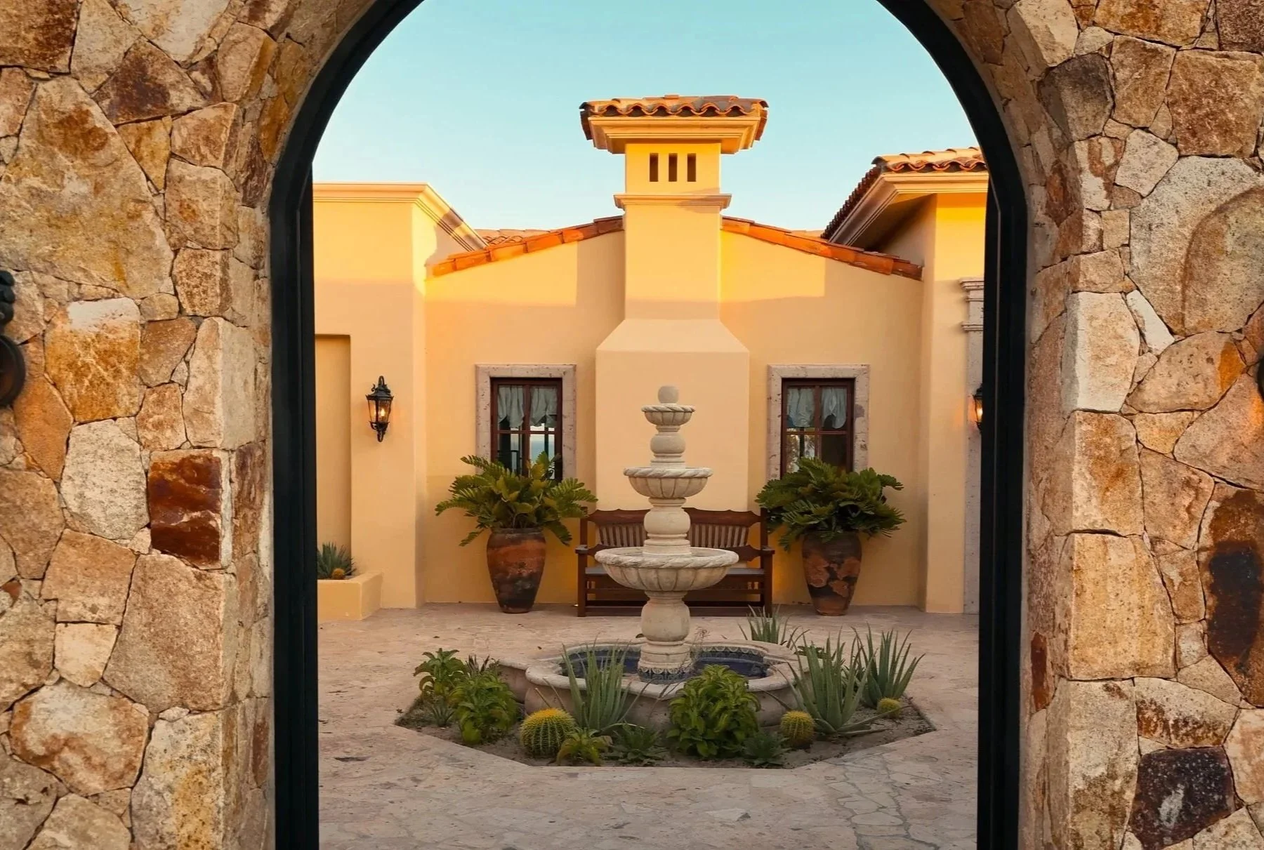 A courtyard view through a stone archway showing a cream-colored house with a terracotta roof, a fountain in the center, potted plants, and a bench beneath two windows.