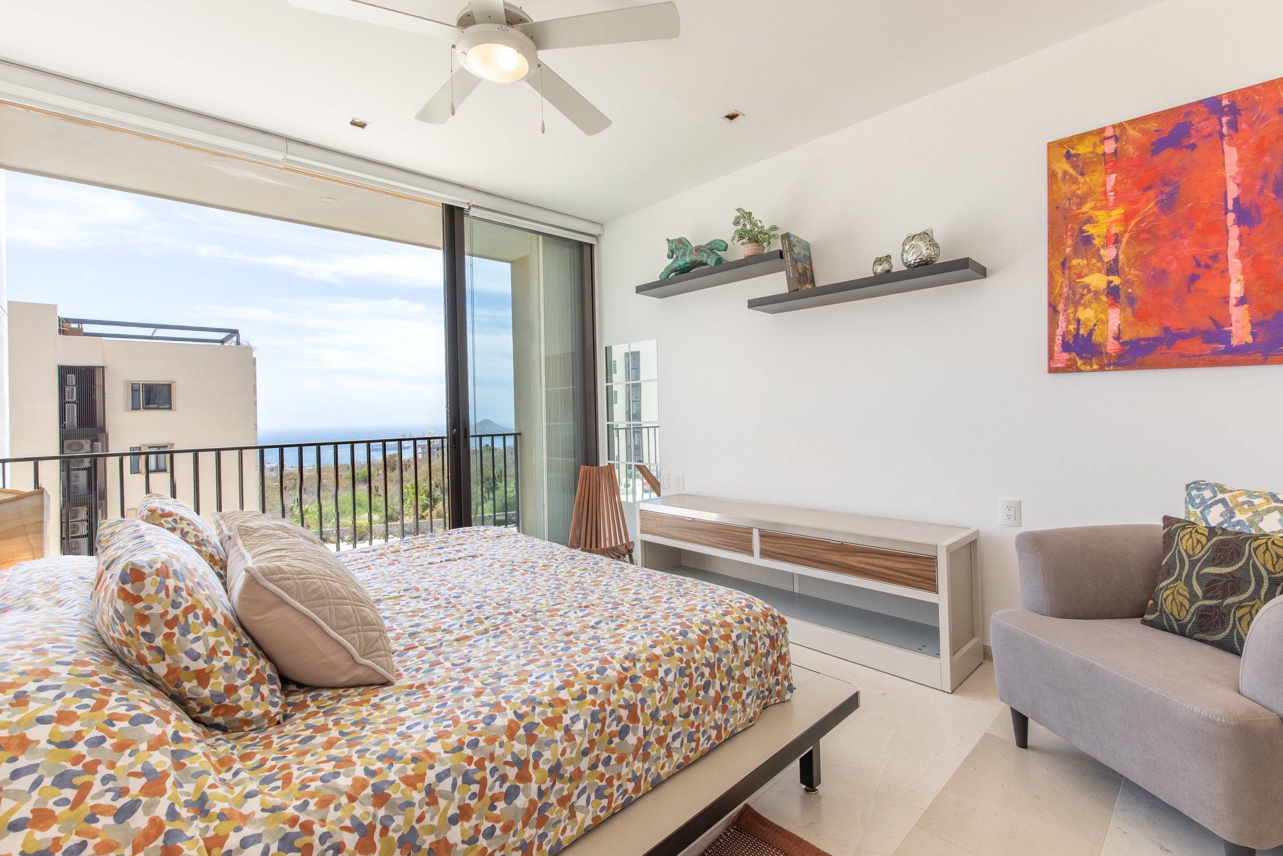 A modern bedroom with a bed covered in a multicolored abstract patterned duvet and matching pillows, a balcony view with buildings and sky outside, floating shelves with decorative items including a sculpture and plants, a vibrant abstract painting o