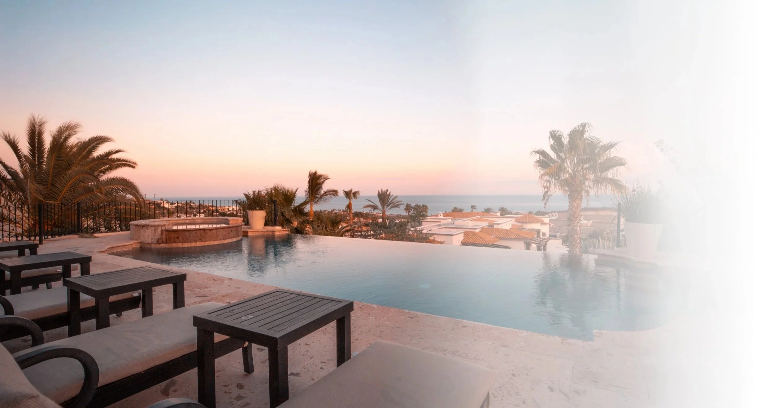 A luxurious outdoor swimming pool area overlooking a coastal town with rooftops and palm trees during sunset or sunrise. San Jose Del Cabo