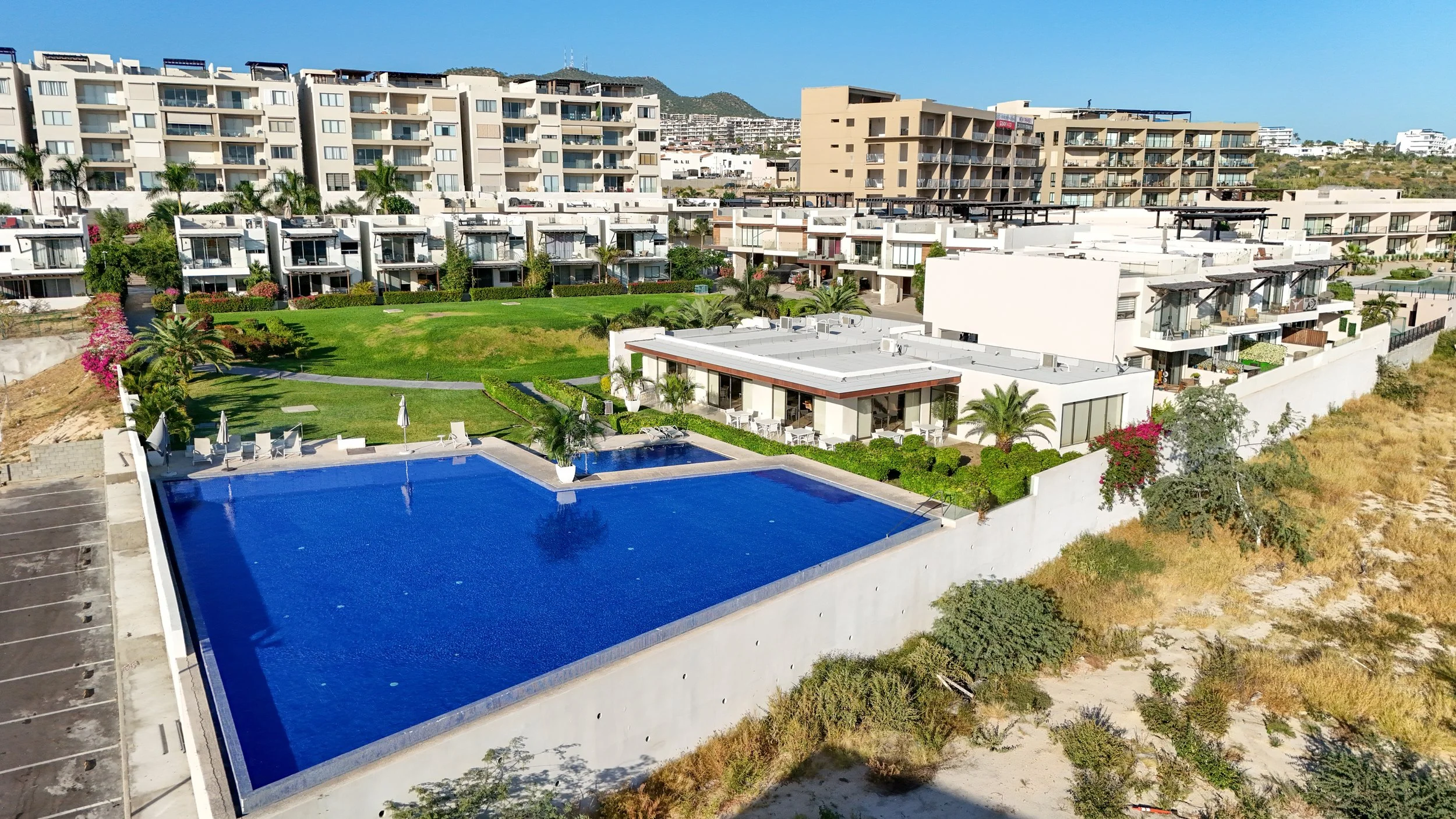 Aerial view of a beachfront residential complex featuring a large swimming pool, green lawn, tropical plants, and multiple modern apartment buildings with balconies, set against a background of hills and a clear blue sky.