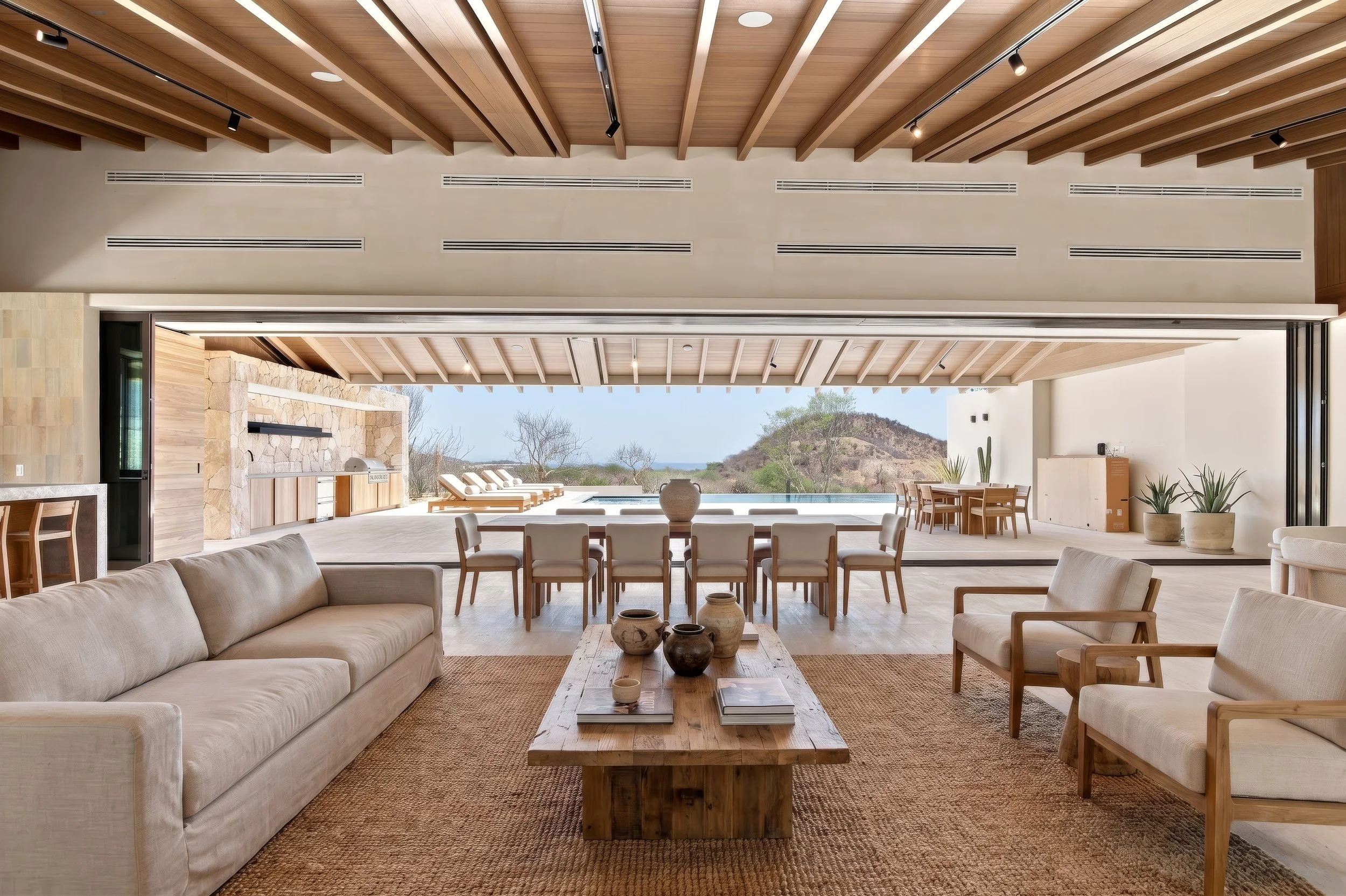 One and Only Palmilla Modern living room with beige sofas, wooden coffee table with decorative vases, and an open view to an outdoor patio area with lounge chairs and scenic hilly landscape.