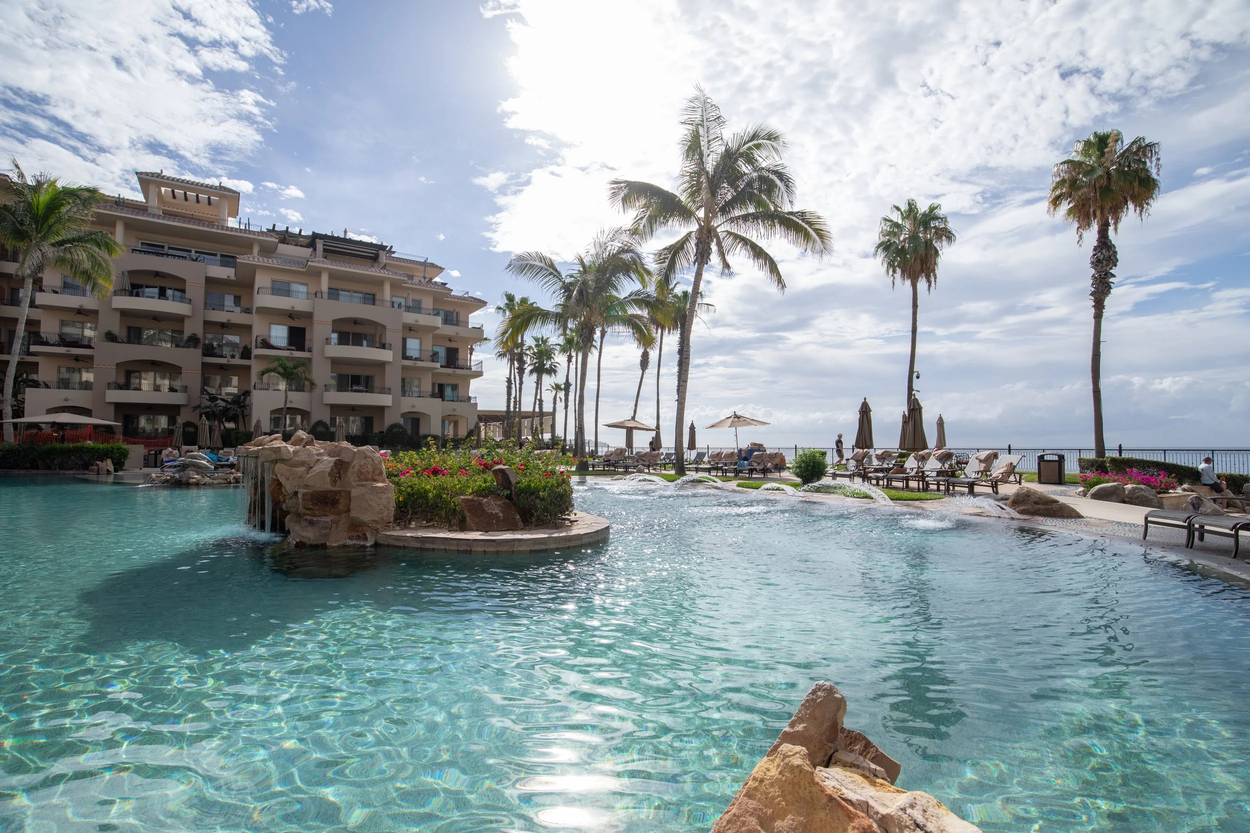 A resort pool area with clear swimming pool water, surrounded by palm trees, lounge chairs with umbrellas, a building with balconies, and a view of the ocean under a partly cloudy sky.