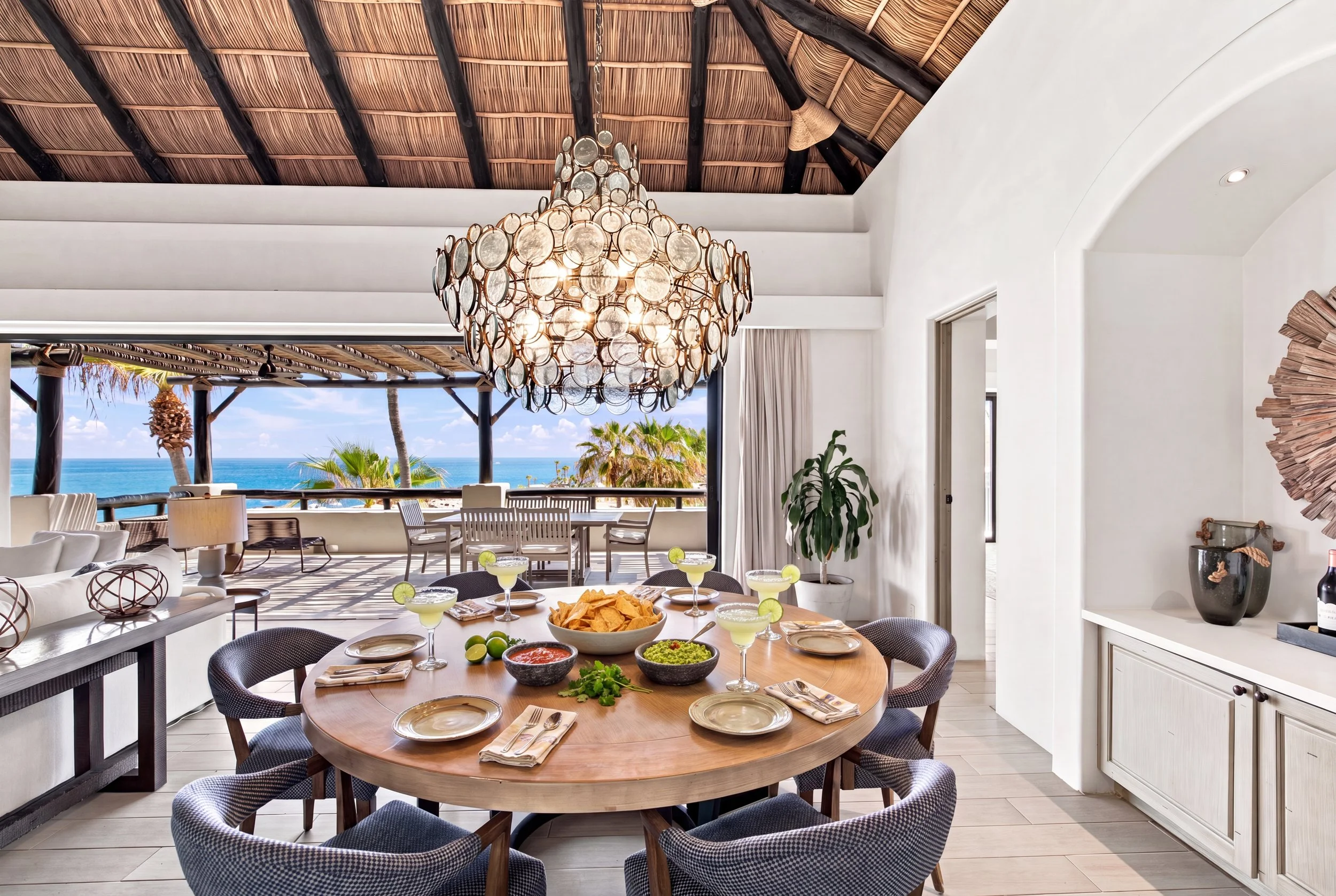 Interior of a room with a round dining table set with food and drinks, a large ornate chandelier hanging from a wooden ceiling, and an open view of an outdoor patio with a view of the ocean and palm trees.
