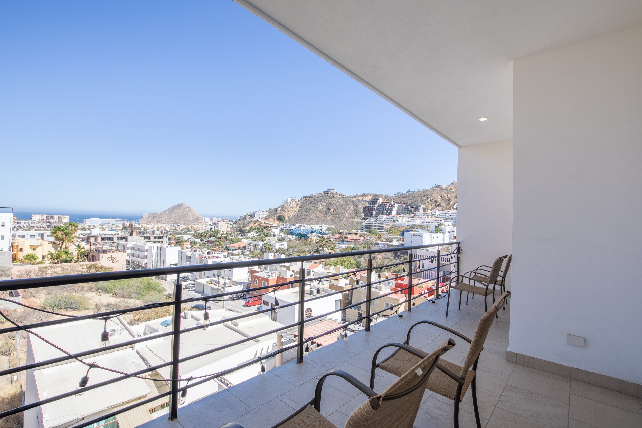 Balcony view overlooking a city with mountains and the ocean in the background, with chairs along the railing.