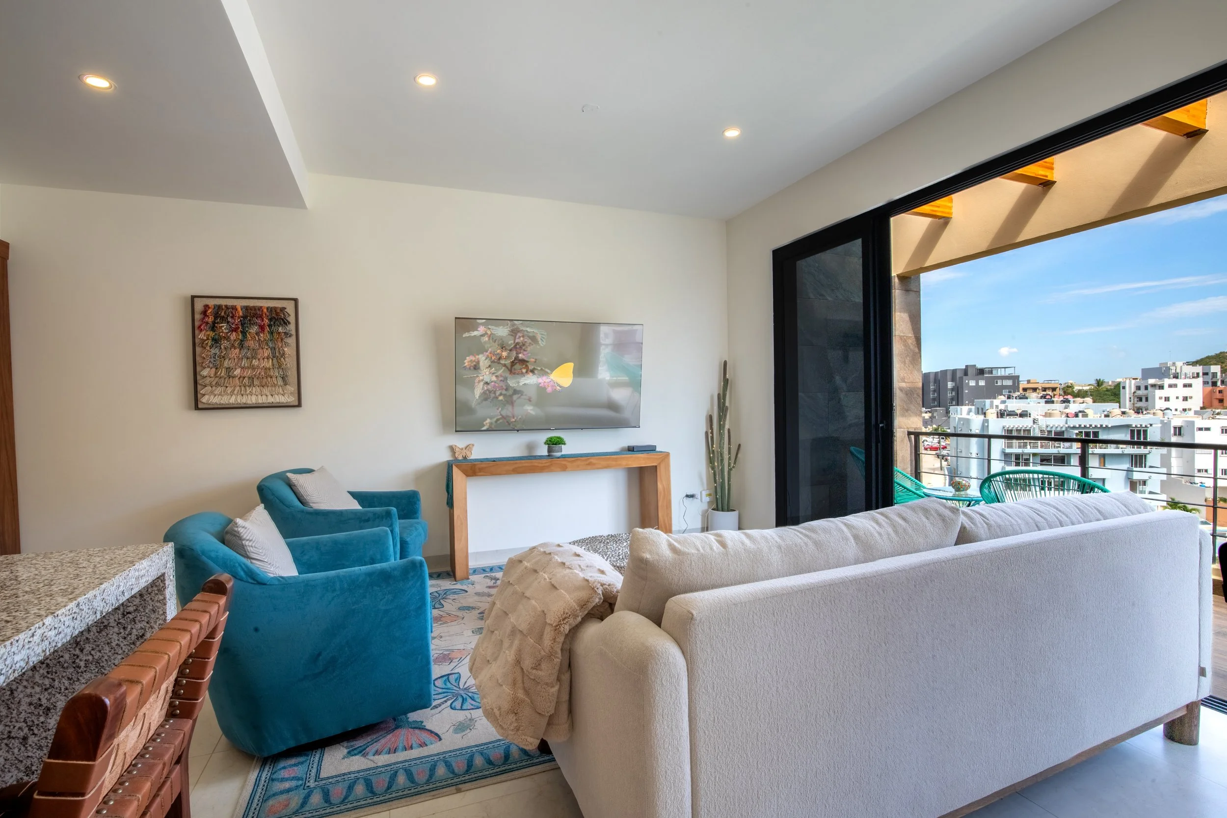 Living room in Montemar with a beige sofa, blue armchairs, wall art, flat-screen TV, a tall cactus, and sliding glass doors leading to a balcony with city view.