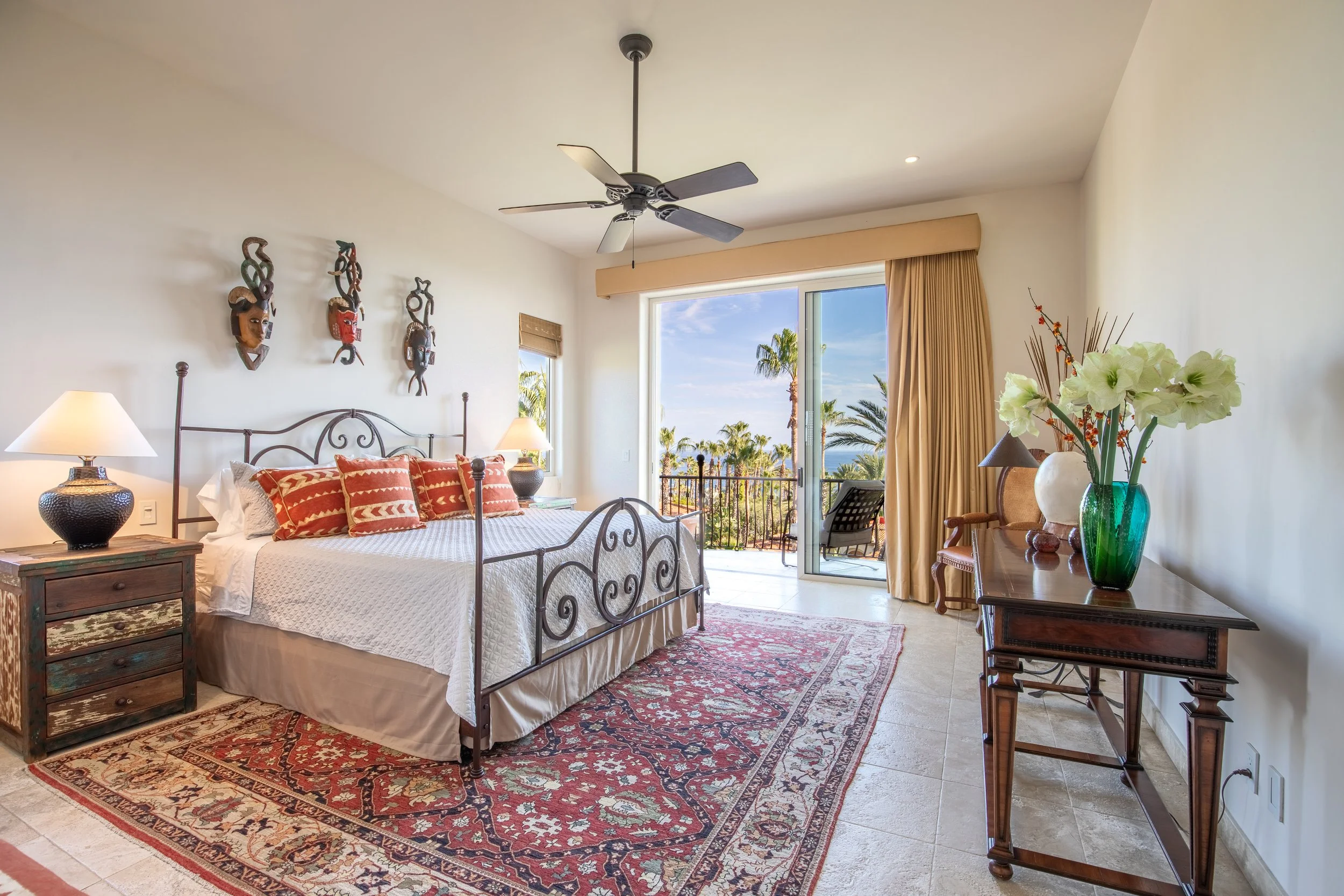 A bedroom with a decorative iron bed, red and white pillows, bedside tables with lamps, a dresser with a large dark vase and white flowers, an armchair, a sliding glass door leading to a balcony, and tropical trees outside.