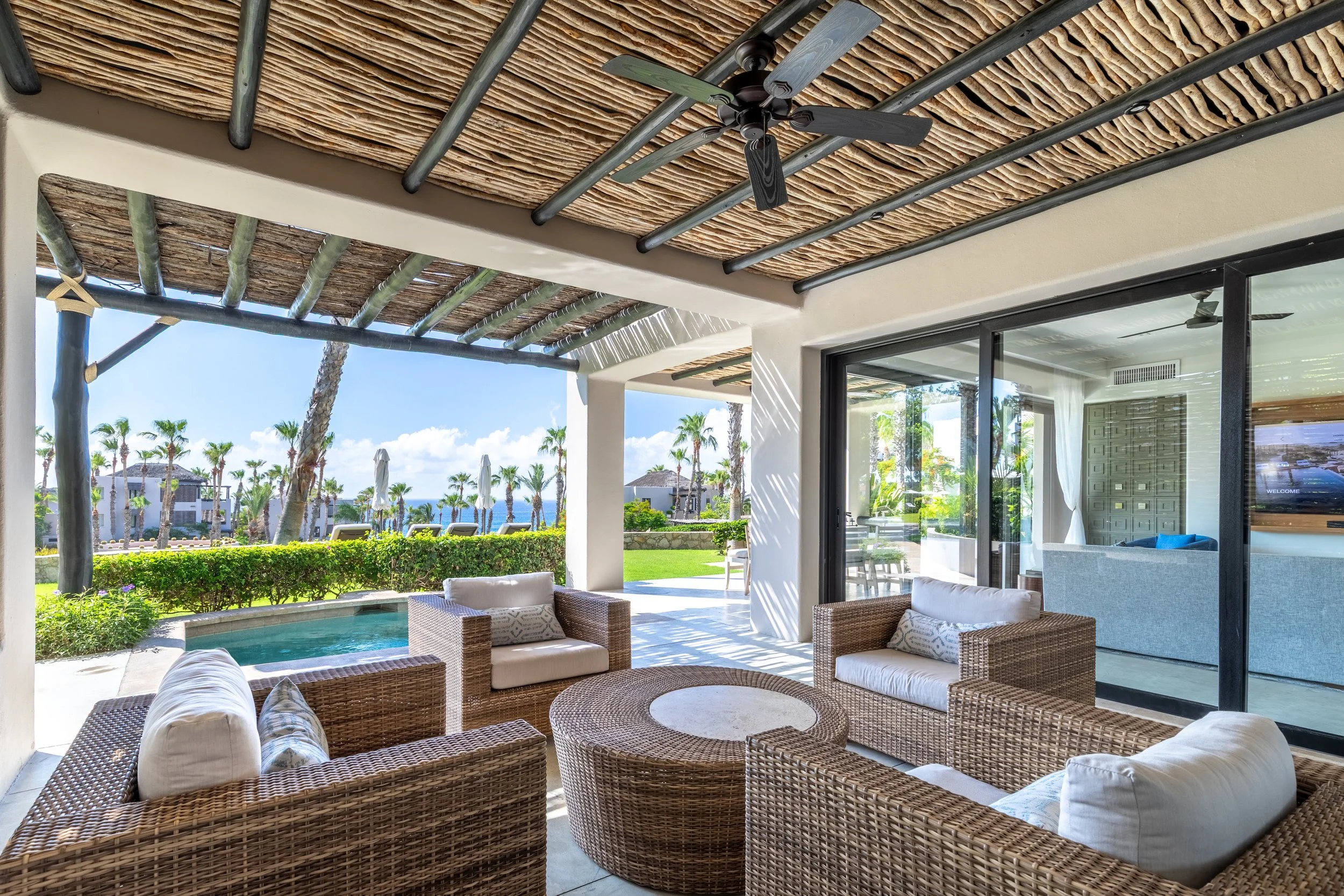 Outdoor patio area with wicker furniture, sliding glass doors, ceiling fans, and a covered thatched roof, overlooking palm trees and a pool with lounge chairs.