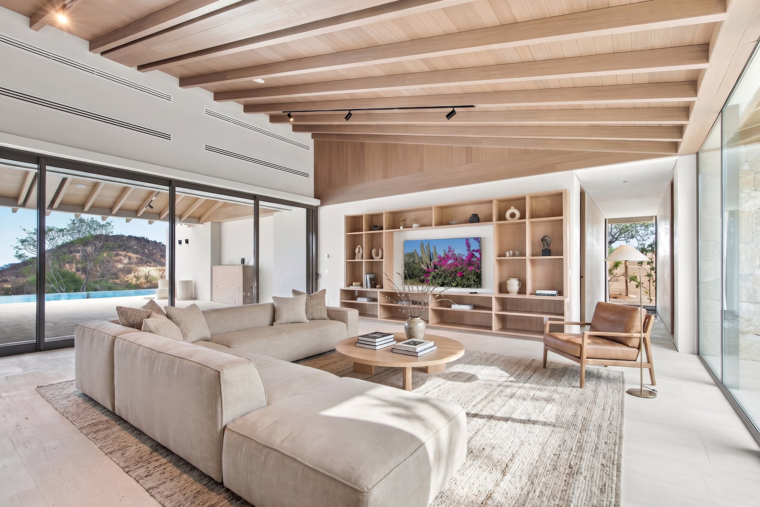 Inside view of a modern living room with a beige sectional sofa, wooden coffee table, and a large built-in wooden entertainment unit with decorative items and a flat-screen television. Large glass sliding doors reveal an outdoor patio and scenic land