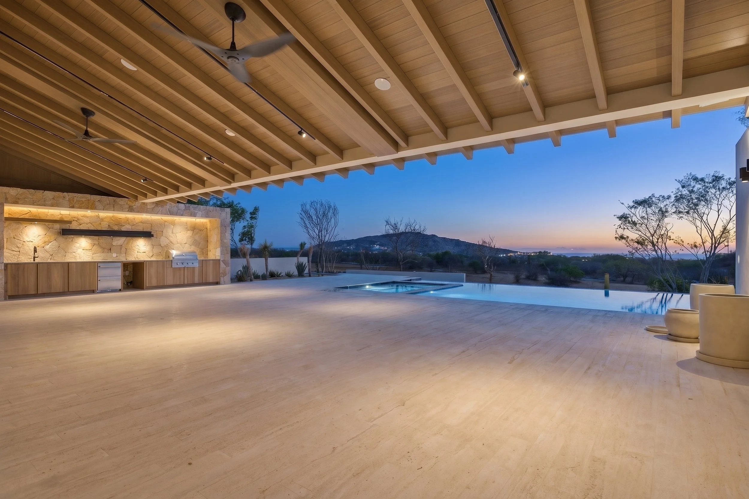 Modern outdoor patio with a roof, ceiling fans, and a view of trees and hills at sunset, featuring a built-in grill and seating area. Luxury real estate photographer.