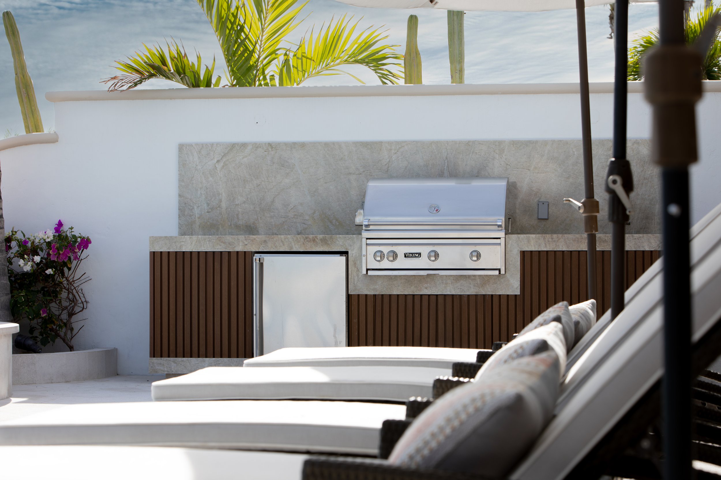 Outdoor patio area with a built-in grill, lounge chairs, and lush greenery in the background.
