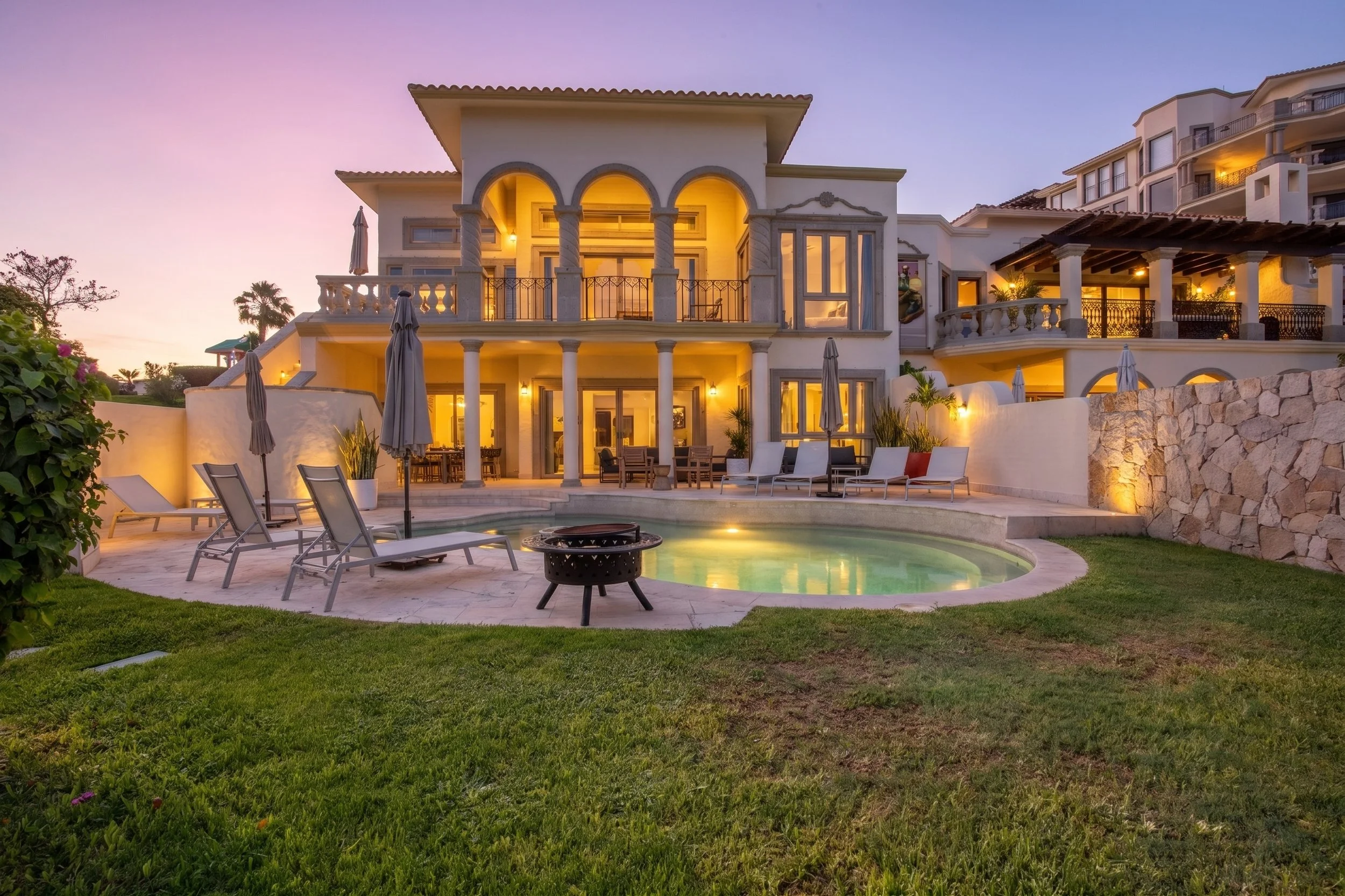 Luxury multi-story house with a swimming pool in front, patio furniture, and illuminated windows at sunset