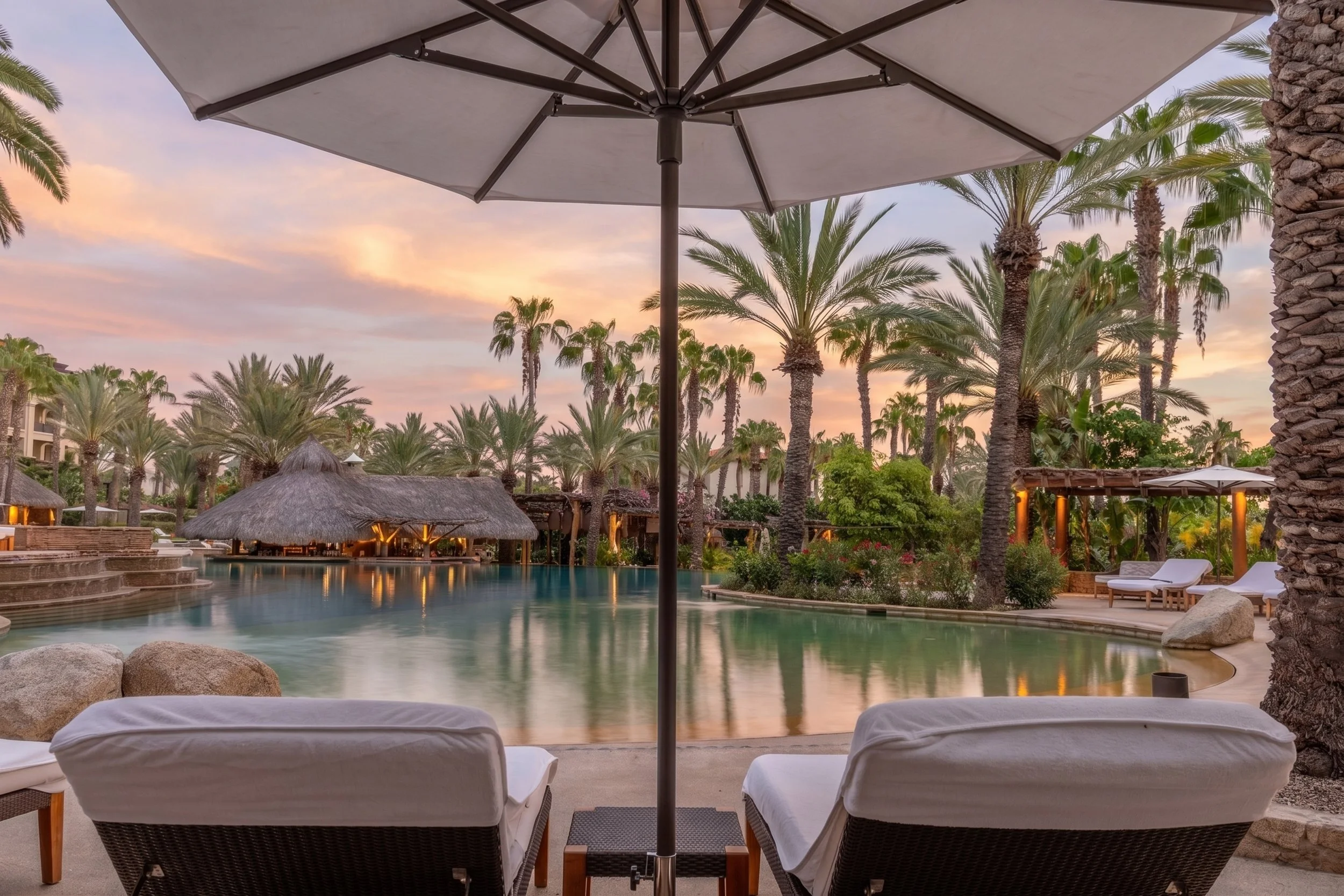 View of a resort pool area at sunset, with lounge chairs under an umbrella in the foreground, surrounded by palm trees and tropical vegetation.