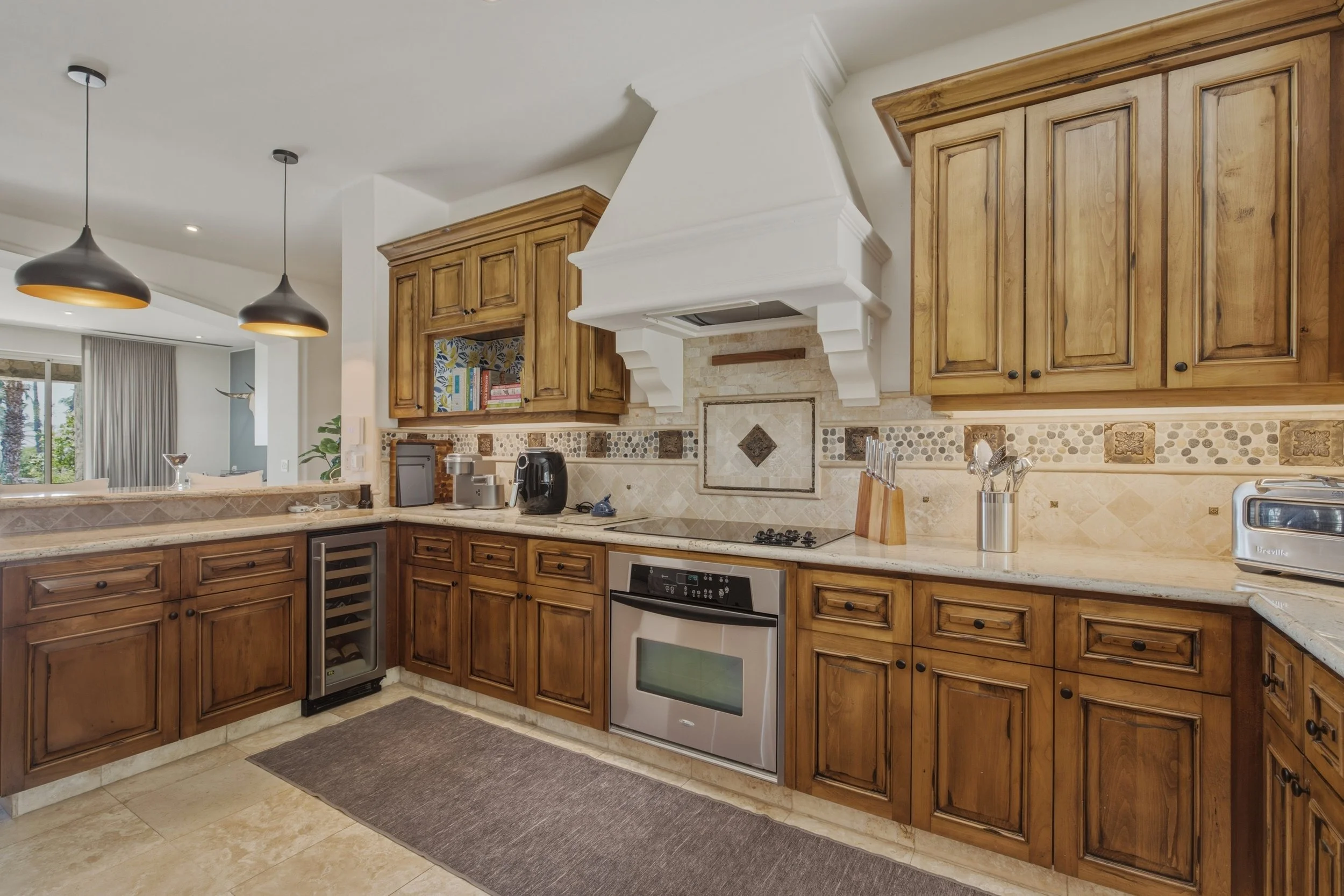 A kitchen with wood cabinets, a white range hood, beige tile backsplash, and black pendant lights hanging from the ceiling.