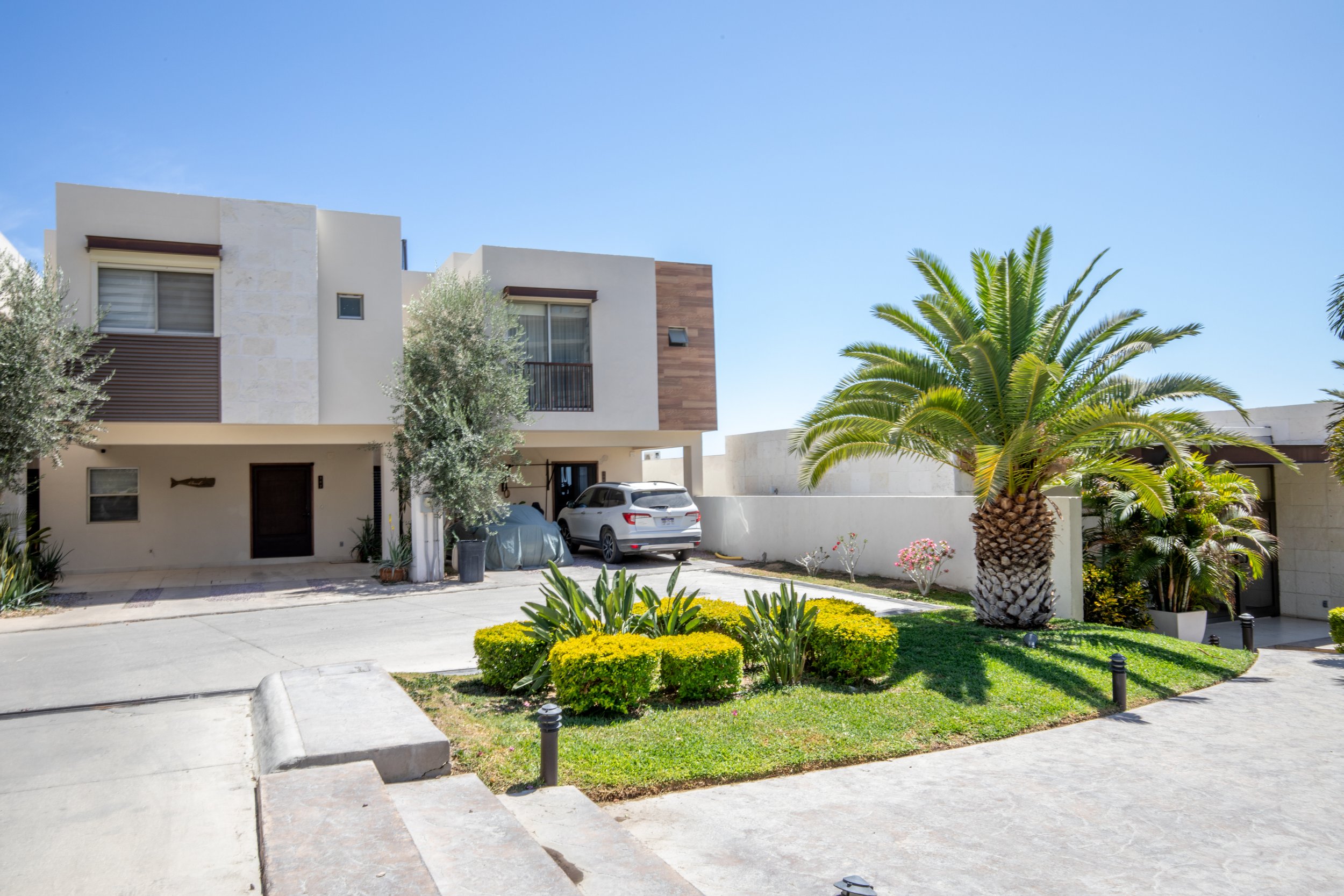Modern residential building with a white facade, a driveway with parked cars, and landscaped greenery including a large palm tree and neatly trimmed bushes under a clear blue sky.