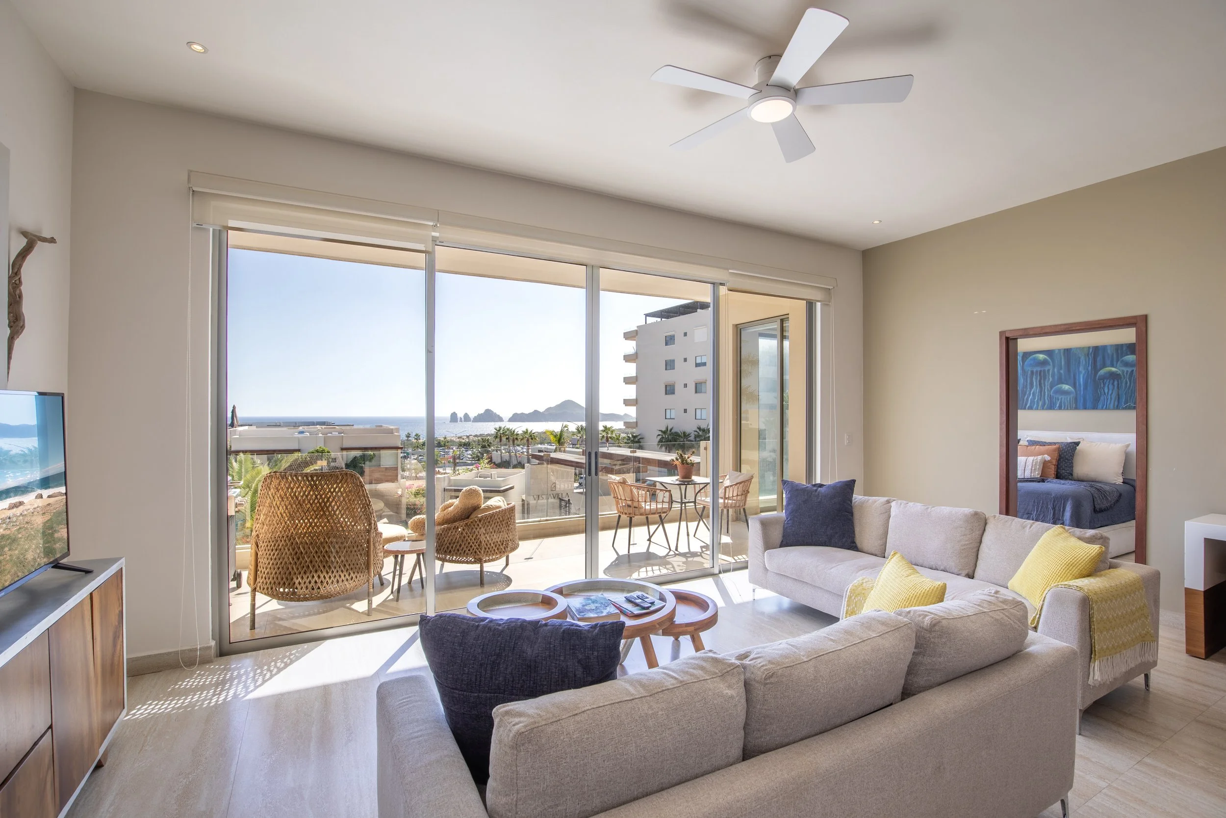 Living room with large sliding glass doors leading to a balcony, ocean view, beige couches with blue and yellow pillows, small wooden tables, TV on stand, ceiling fan, bedroom visible through mirror, sunny and bright atmosphere.