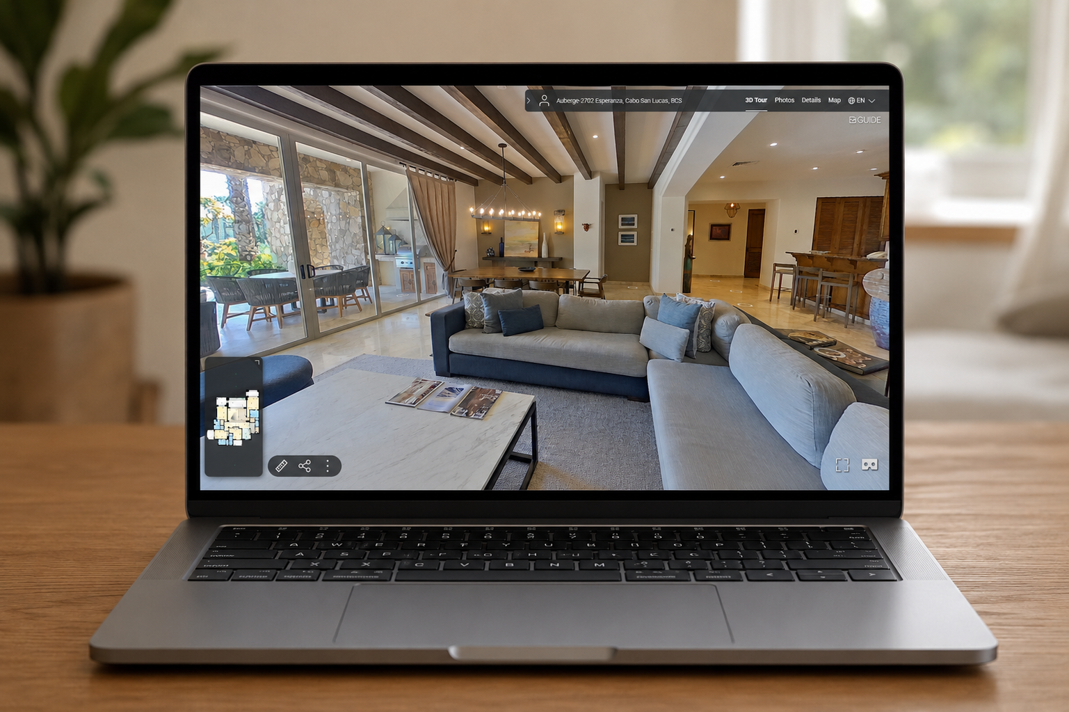 Laptop on a wooden table displaying a photo of a modern living room with a beige and navy sofa, a round coffee table, a dining area with a chandelier, and sliding glass doors leading to an outdoor patio.