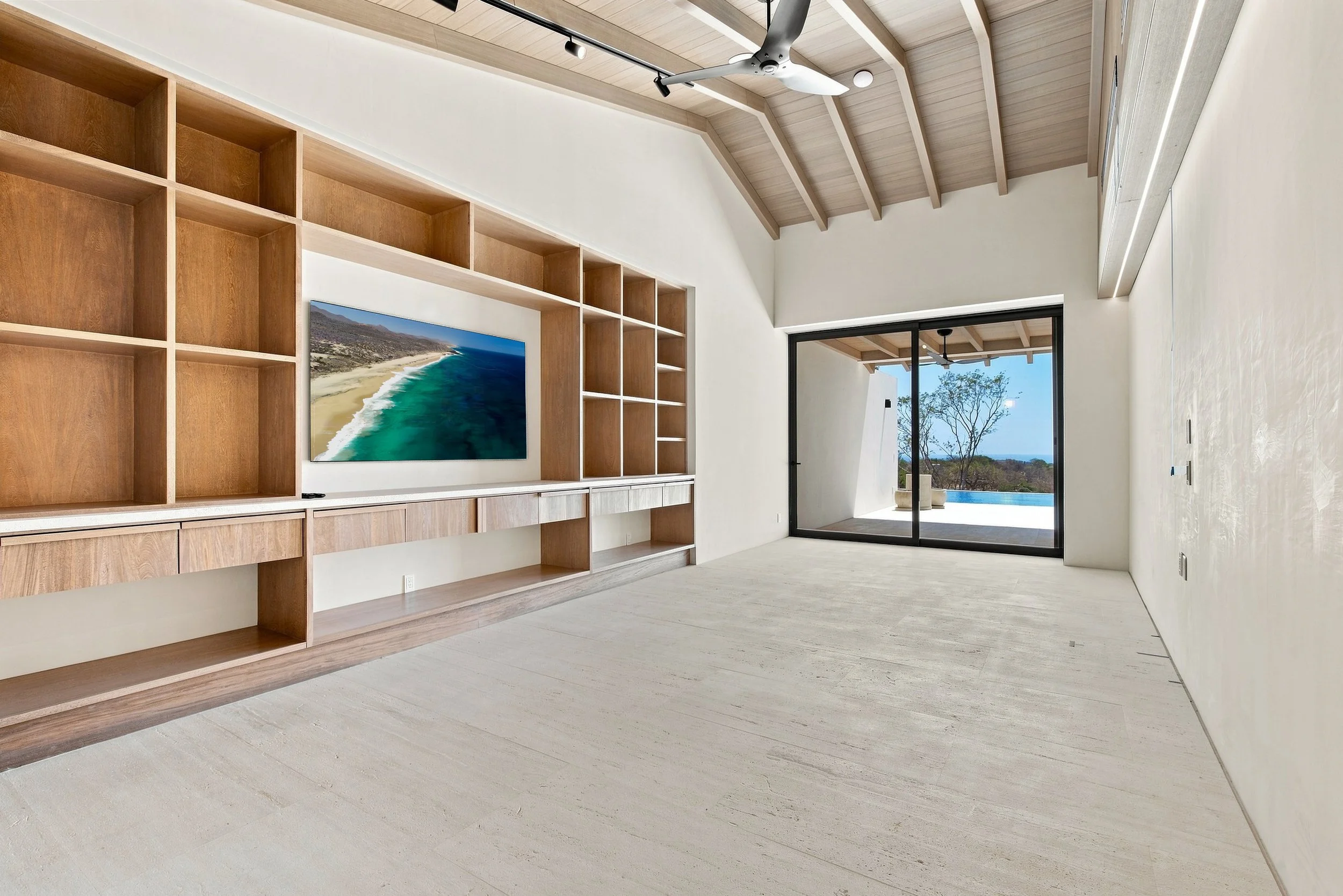 Empty modern living room with built-in wooden shelves, a wall-mounted TV displaying a coastline, large glass sliding doors opening to a patio with trees and ocean view, and a high wooden ceiling with beam details. Custom build in San Jose Del Cabo.