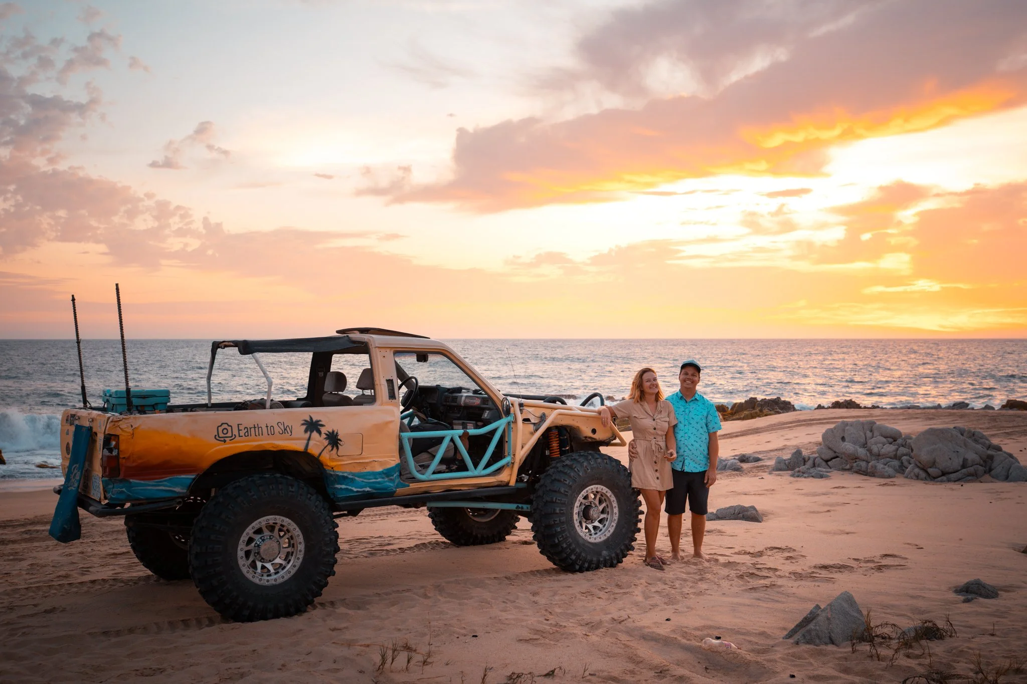 Two smiling people, a woman and a man, standing on a sandy beach next to a colorful off-road vehicle during sunset. The vehicle has a tropical-themed design with palm trees and the words "Earth to Sky" on the side, and there are rocks and the ocean in the background.