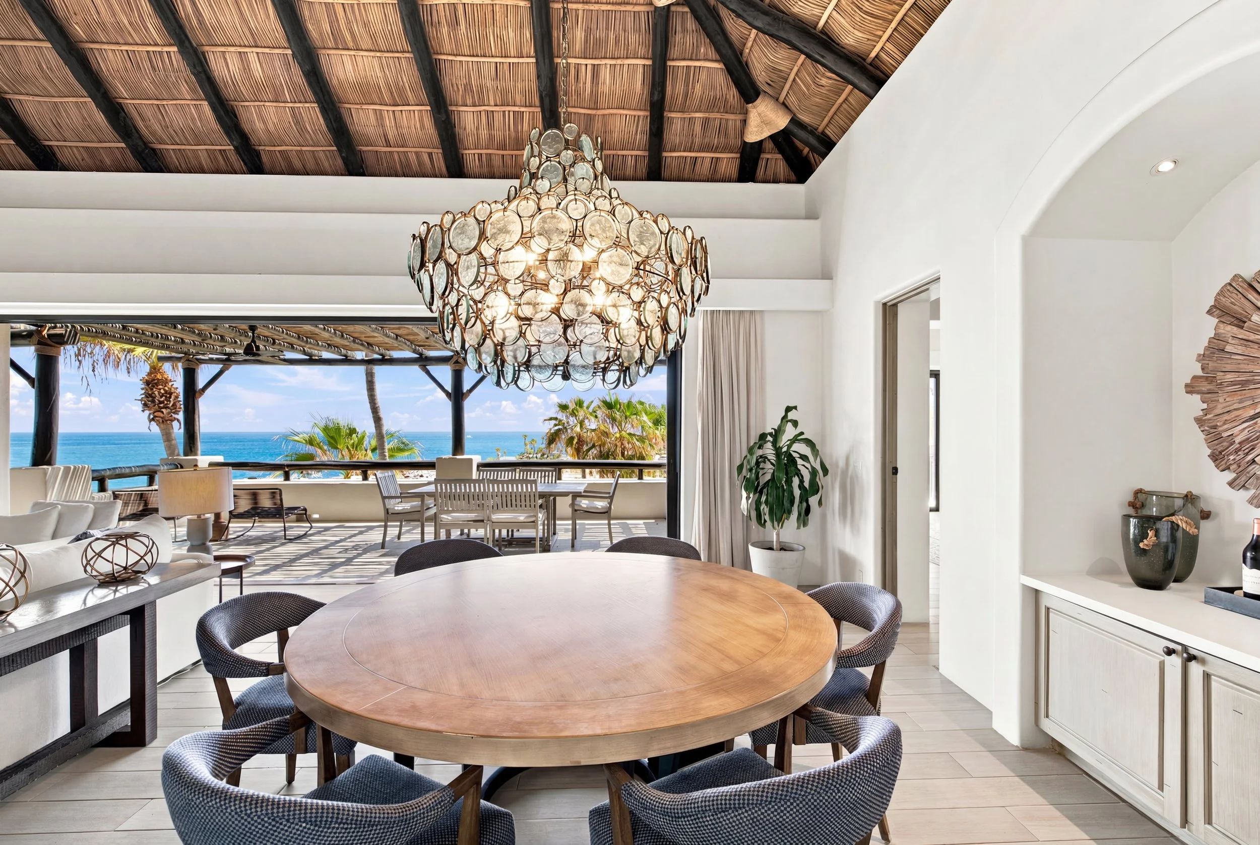Interior view of a bright, open room with a wooden round dining table surrounded by blue patterned chairs. A large chandelier made of circular glass and metal hangs from a thatched, dark wood ceiling. Lounge seating, potted plants, and large windows 