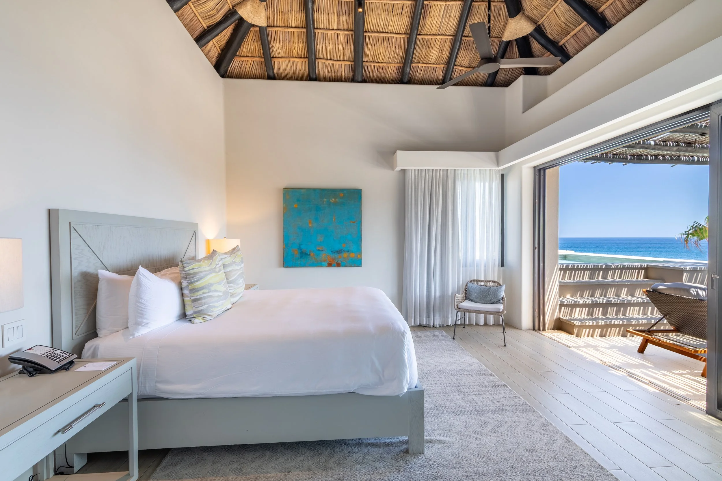 A bedroom with a large bed, white bedding, and pillows, a nightstand with a phone and phone book, a painting on the wall, a ceiling fan, and an open sliding door revealing a balcony with a view of the ocean.