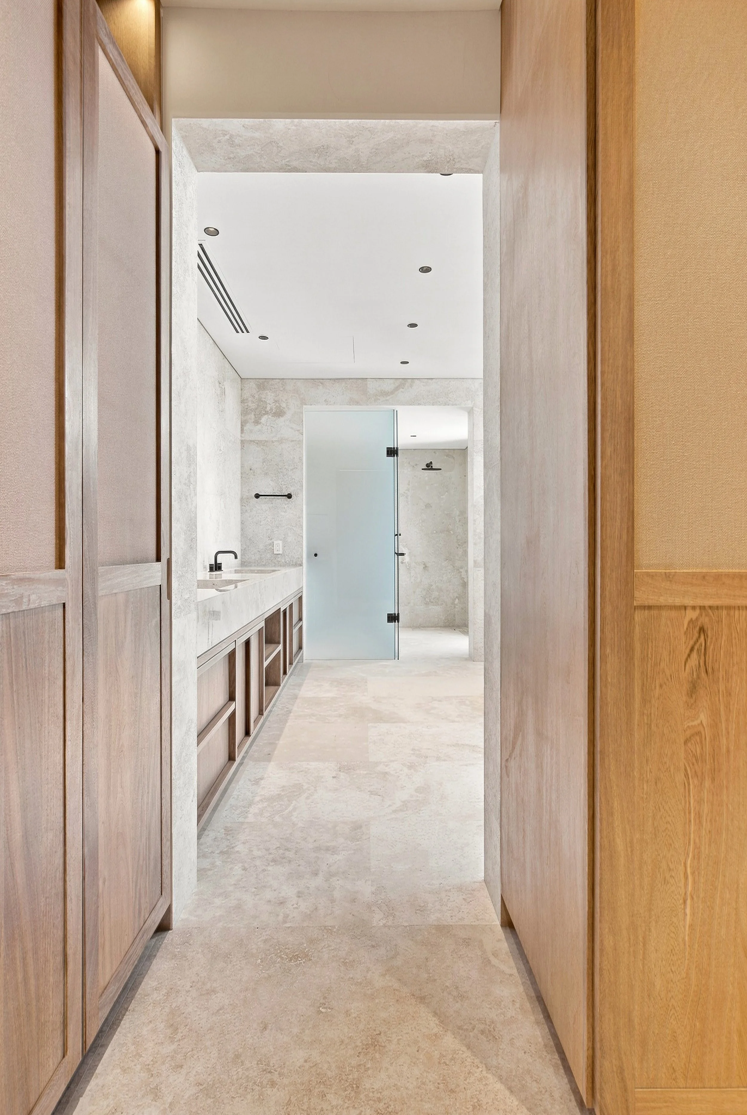 A hallway with beige tiled floors and wood-paneled walls, leading to a bathroom with a frosted glass door, a black towel bar, and a white vessel sink. Custom home in Palmilla