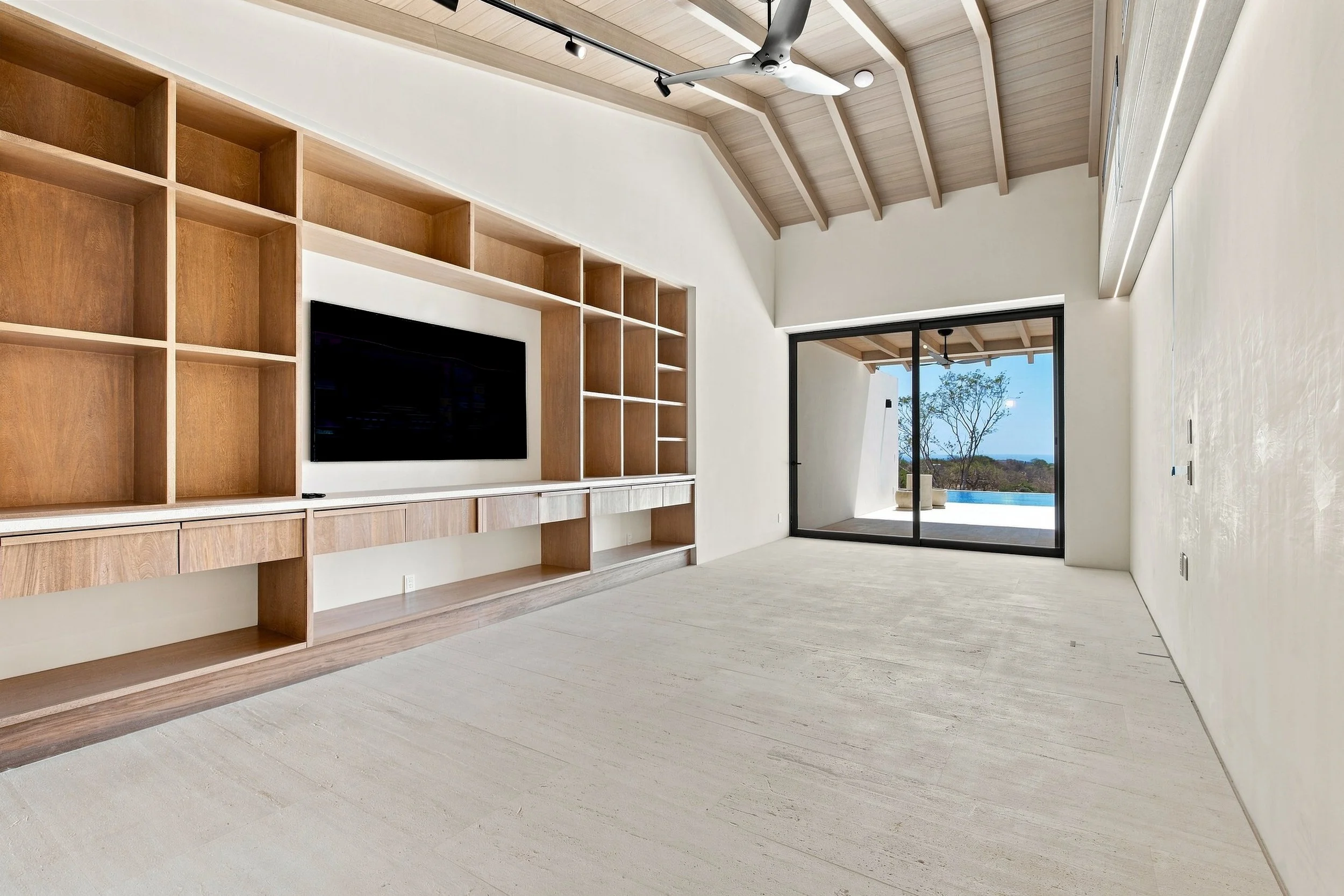 Modern living space with built-in wooden shelves, a mounted flat-screen TV, a ceiling fan, large glass sliding door, and a view of trees outside.