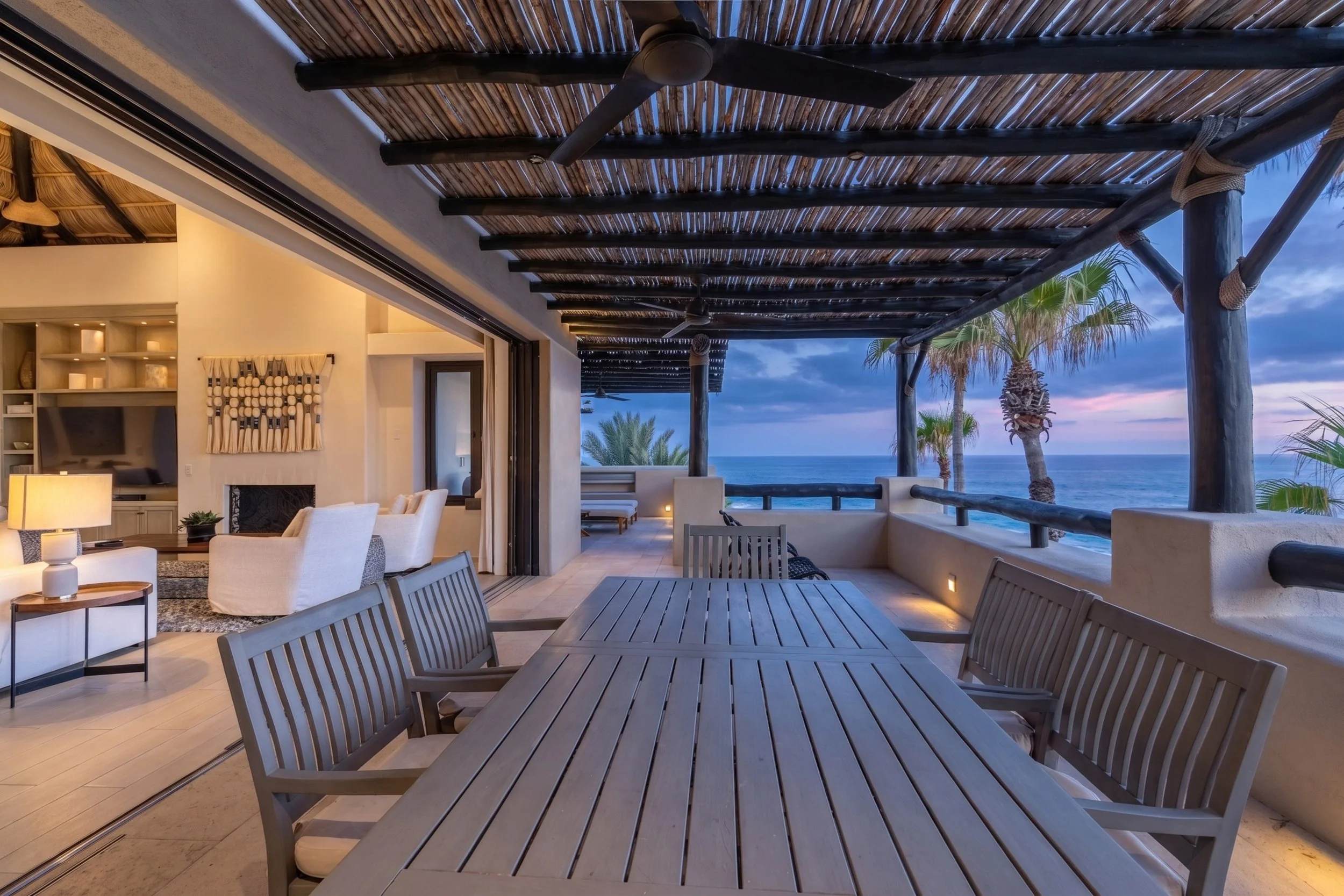 Open-air patio area of a beachside house with wooden dining table and chairs, overlooking the ocean and palm trees at sunset, with a thatched roof and ceiling fans.
