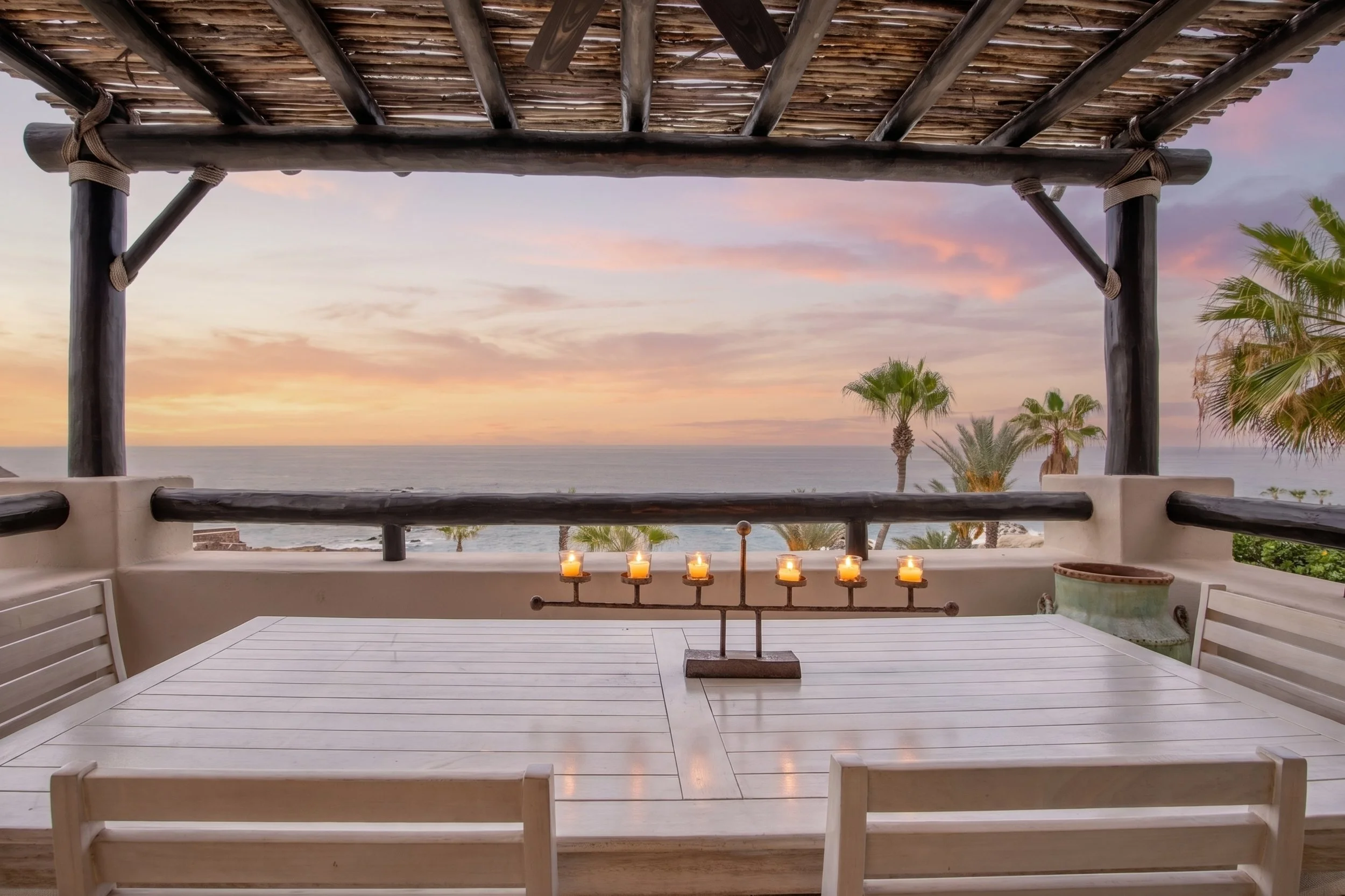 Rooftop terrace with white wooden furniture and candles, overlooking the ocean at sunset with palm trees
