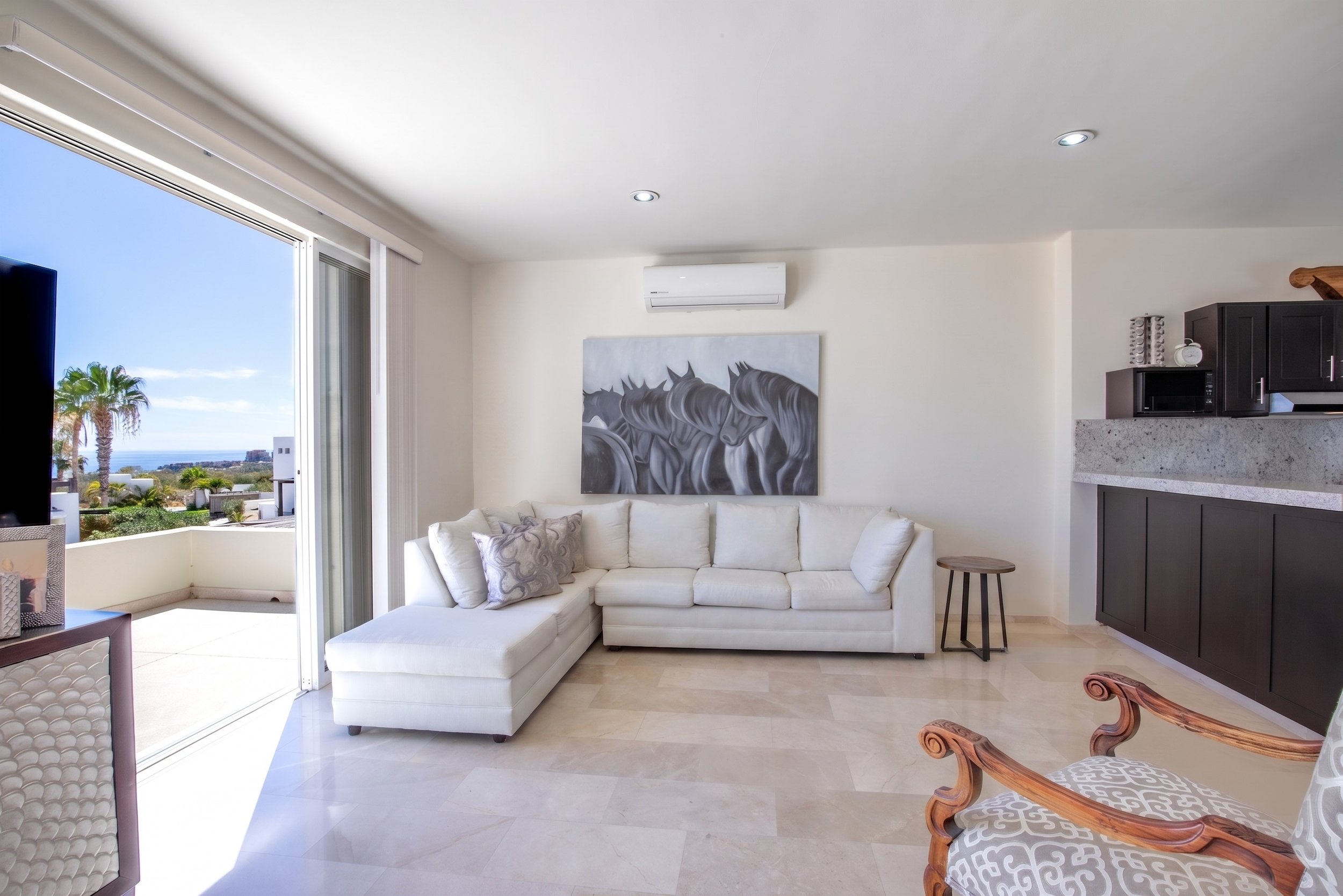 Living room with white sectional sofa, painting of black horses on wall, sliding glass door leading to balcony with outdoor view of palm trees and ocean, black kitchen cabinets, microwave, air conditioning unit, and wooden chairs.