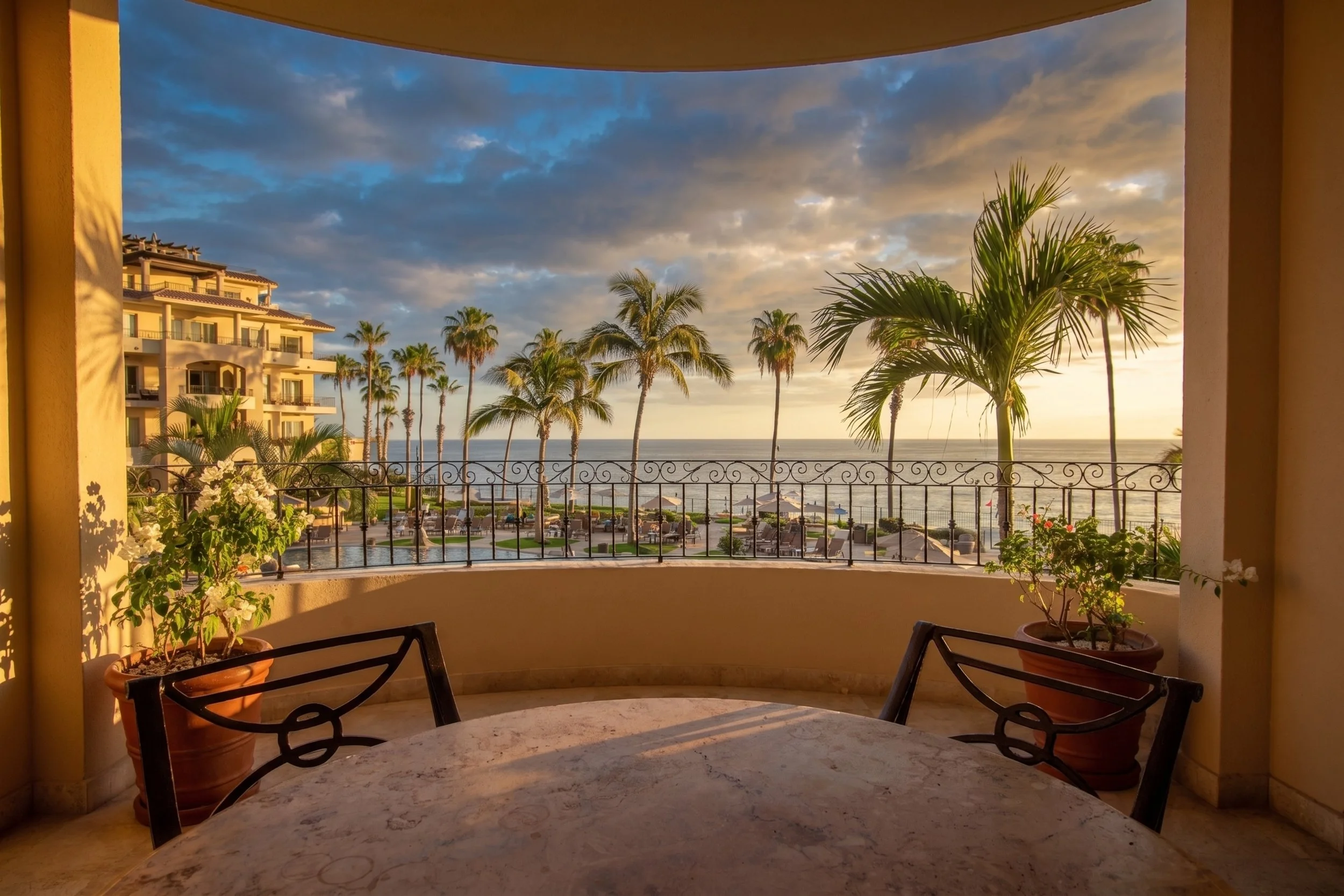View from a balcony showing palm trees, a building with multiple balconies, a swimming pool area, and the ocean in the background under a partly cloudy sky at sunset.