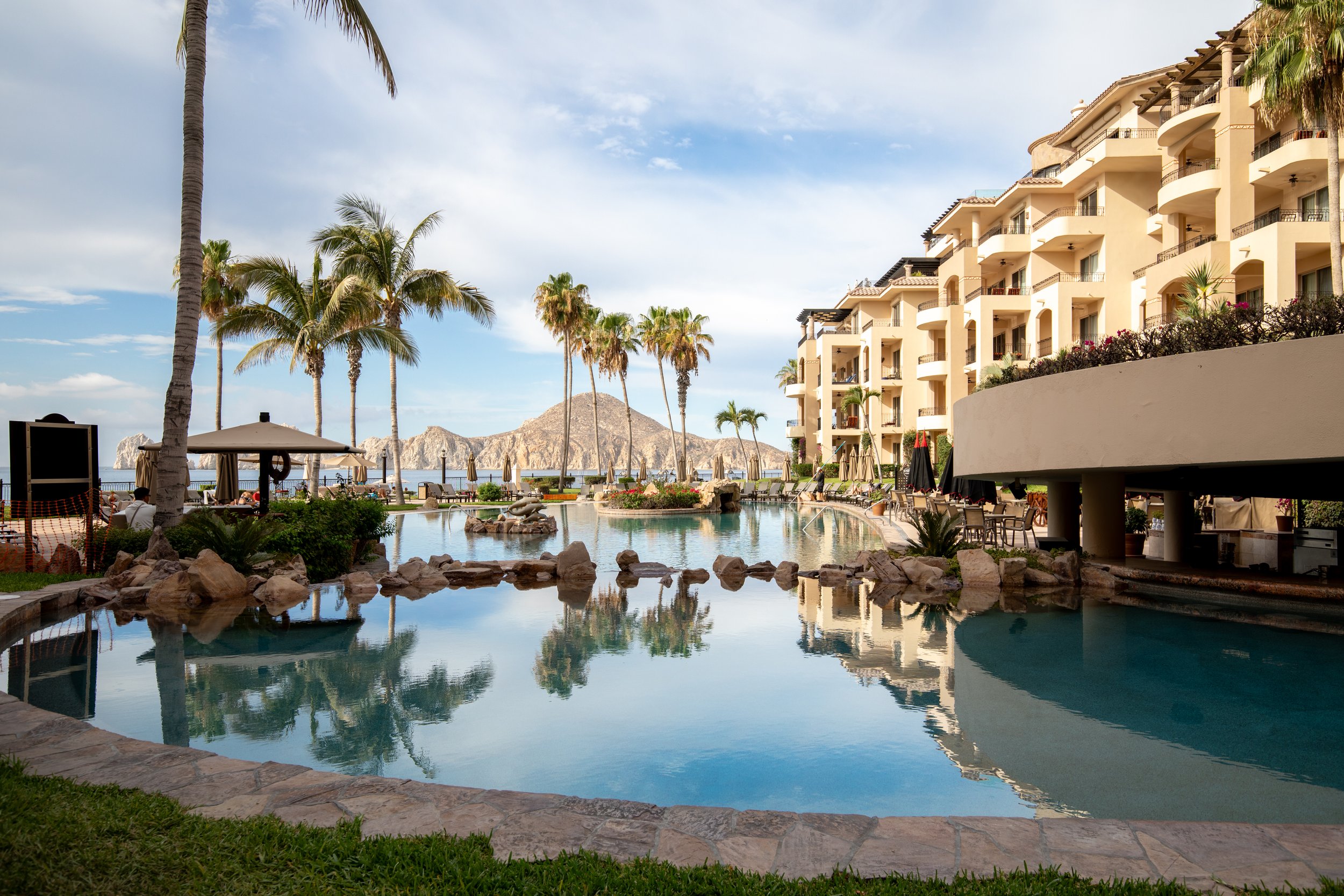 A scenic view of Villa La Estancia resort pool area with palm trees, lounge chairs, umbrellas, and a multi-story building overlooking the ocean and mountains in the distance under a partly cloudy sky.
