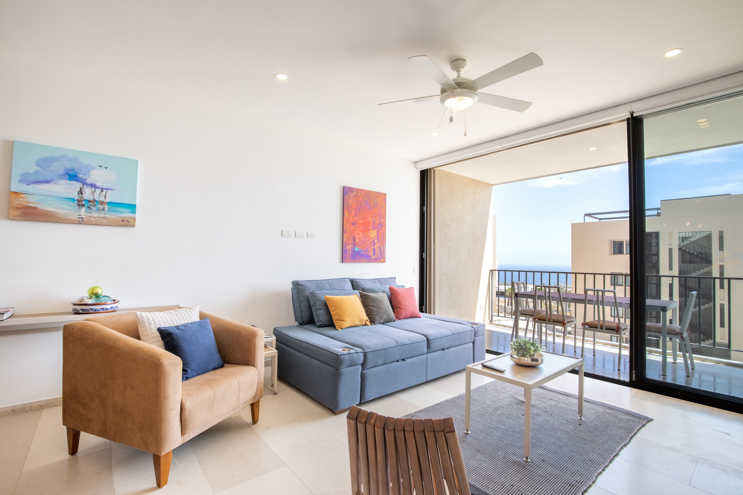 Living room with a beige armchair and a blue sofa, colorful pillows, wall art, small table with a plant, large sliding glass door opening to a balcony with outdoor furniture and ocean view.