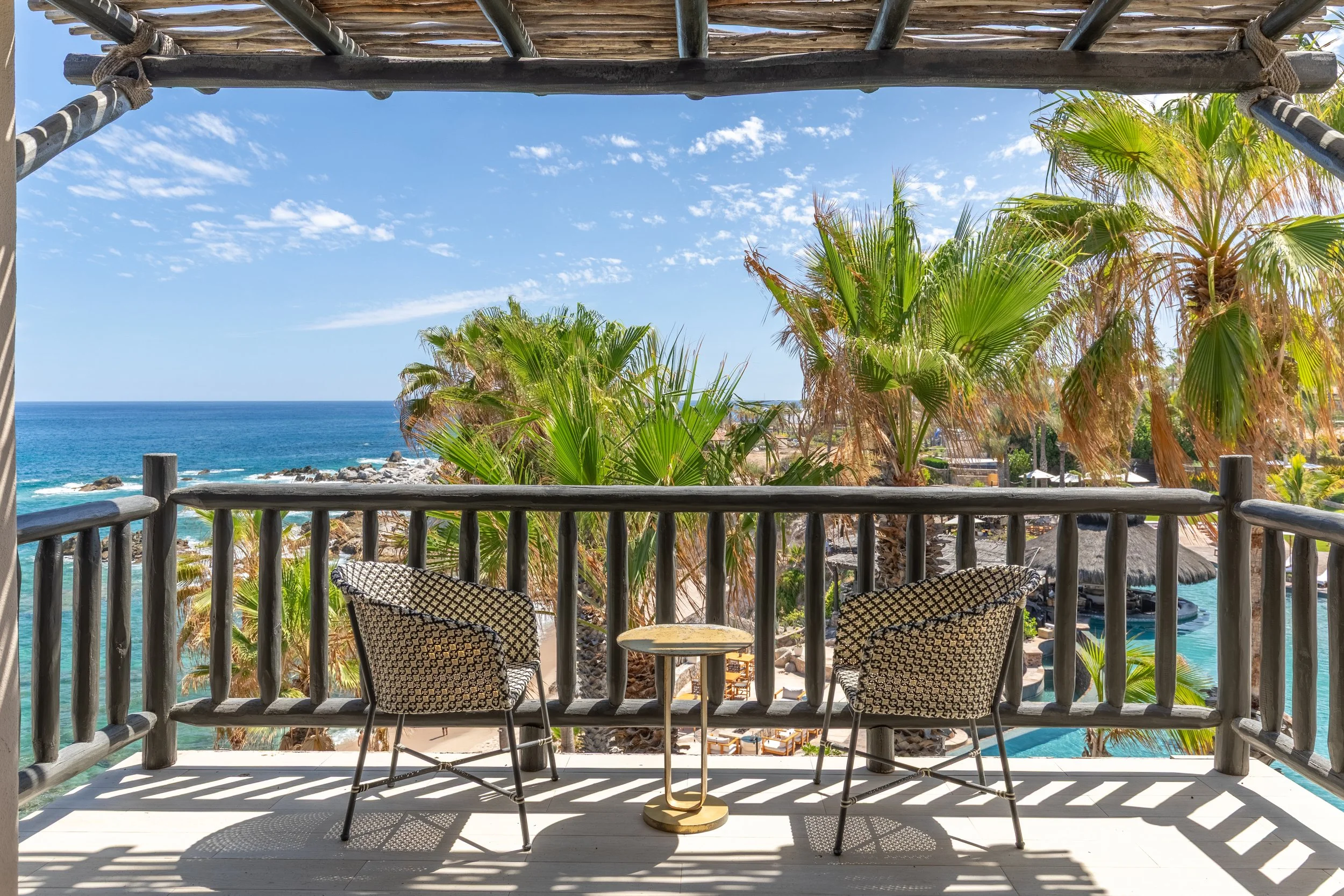 A balcony overlooking Esperanza and the ocean with palm trees, some beach chairs, and an umbrella visible in the distance. The sky is blue with scattered white clouds.