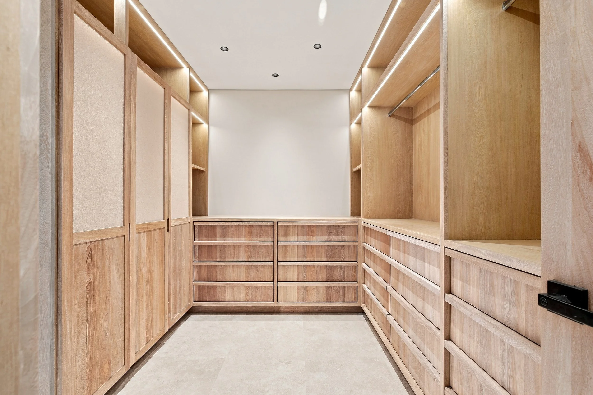 Empty walk-in closet with wooden shelves and drawers on both sides, beige walls, and recessed lighting on the ceiling.