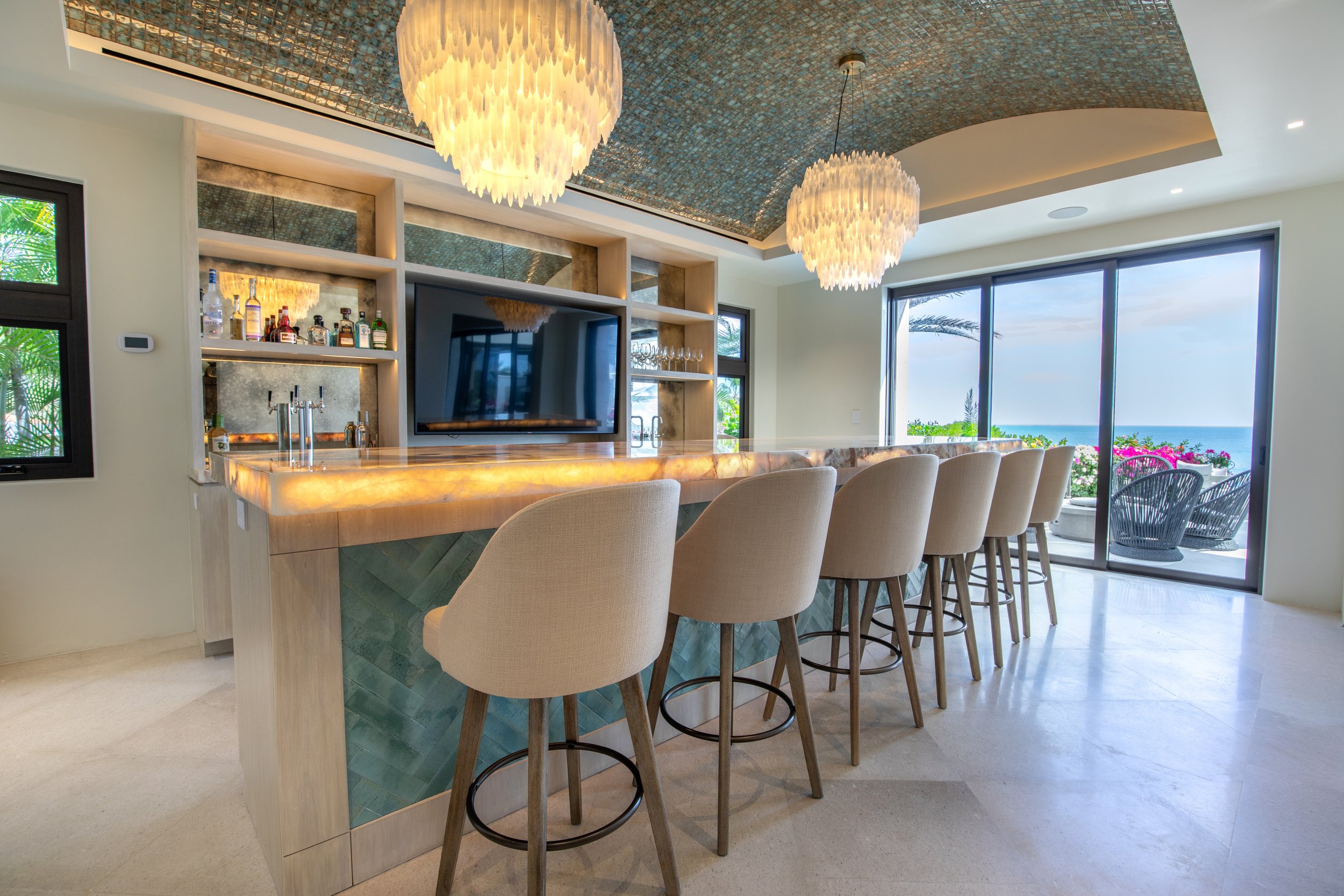 A modern coastal home bar with a large illuminated marble countertop, beige bar stools, a flat-screen TV, and an ocean view through large sliding glass doors with a balcony decorated with pink flowers and outdoor chairs.